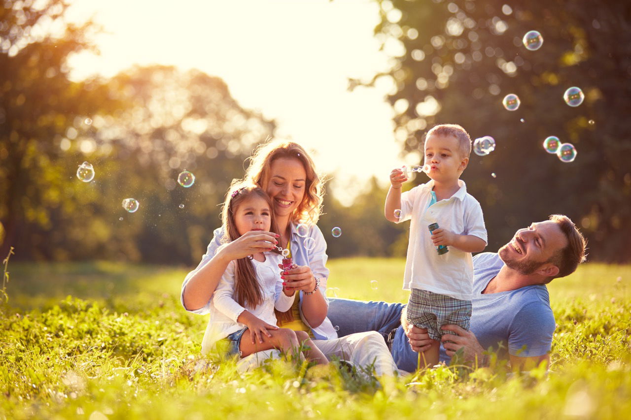 A joyful family of four blowing bubbles and sitting on grass in a sunny park.
