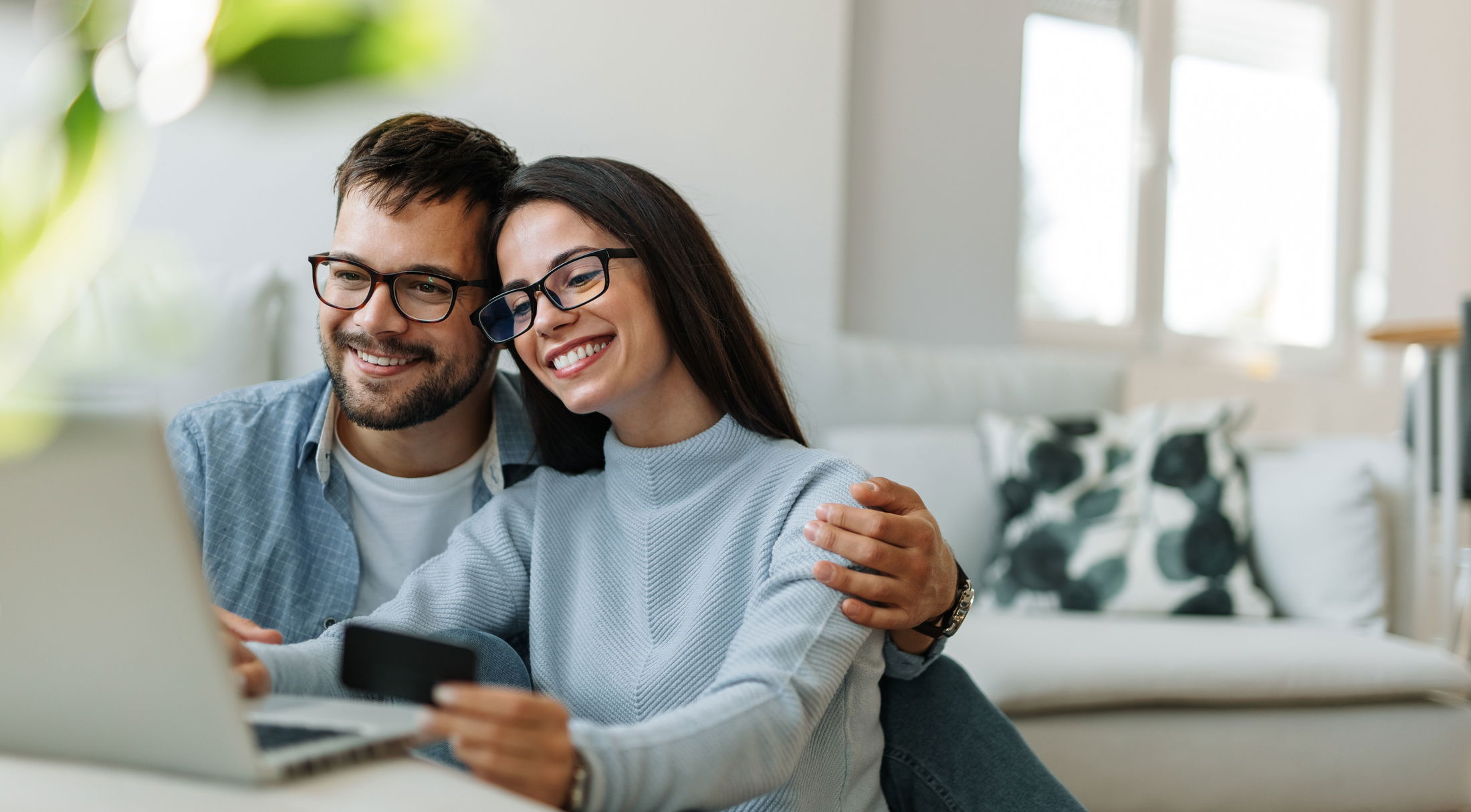 A smiling couple with glasses sits on a couch using a laptop and holding a credit card in a bright, modern living room, illustrating online shopping or remote work.