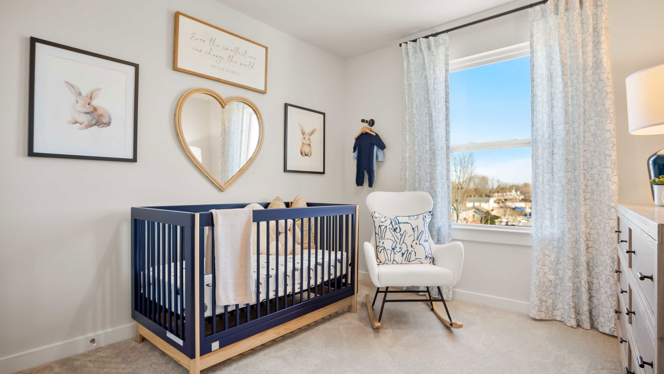 Bright and cozy nursery room with a navy blue crib, heart-shaped mirror, rabbit-themed decor, and a comfortable rocking chair.