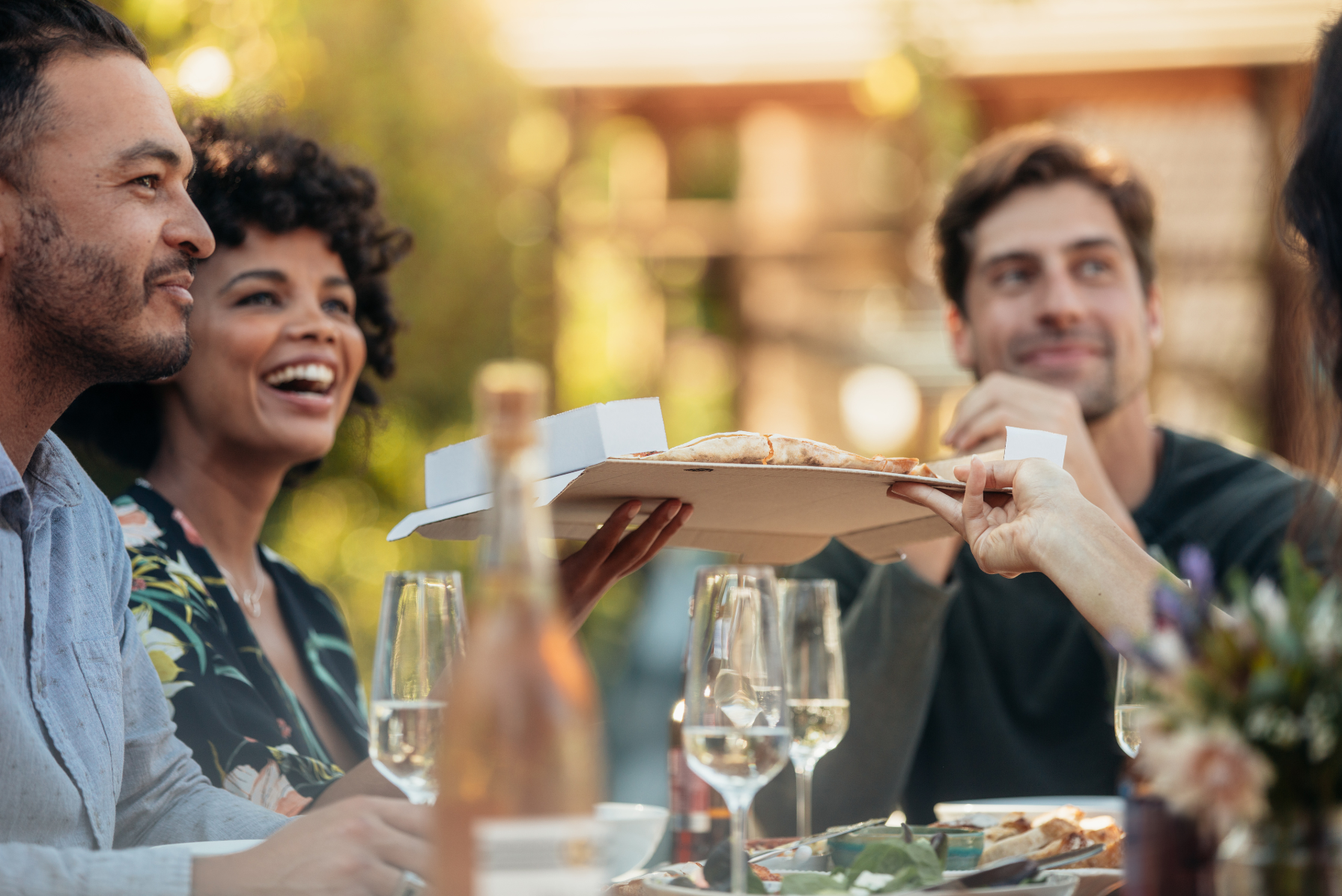 A group of friends enjoys a lively outdoor meal, sharing pizza and wine in a sunny garden setting.
