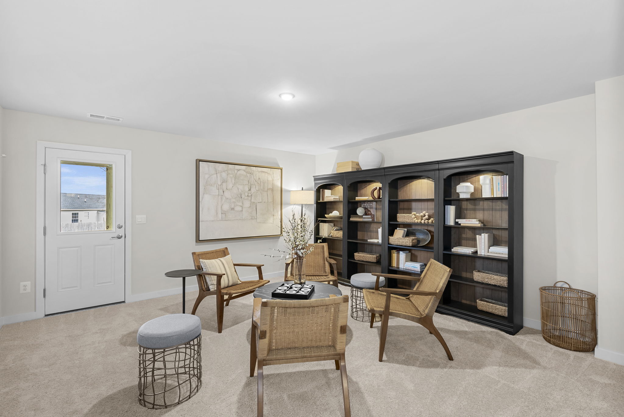 Modern living room featuring wicker chairs, a round coffee table, and a large black bookshelf filled with decor items, set against a neutral-toned carpet and wall art.