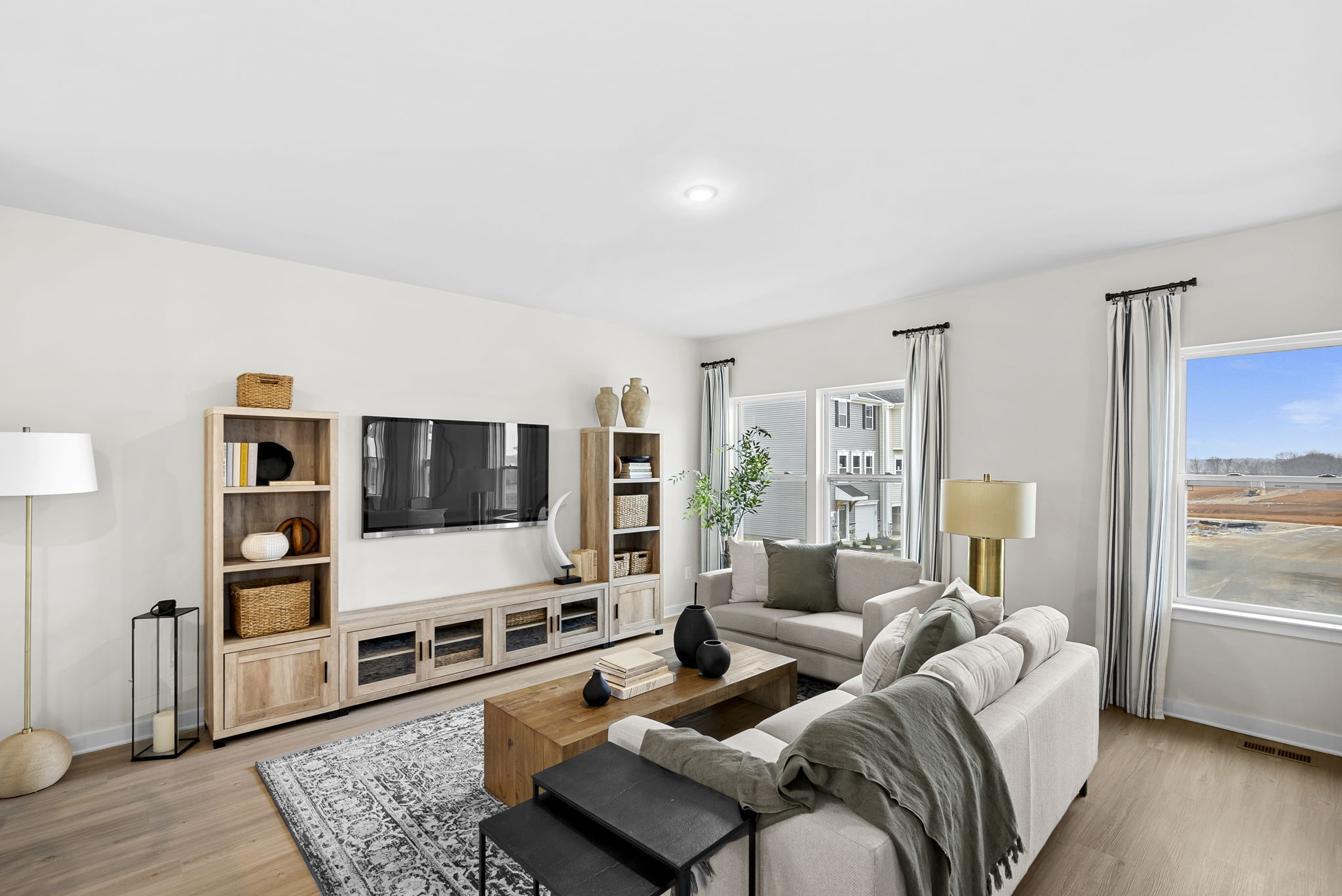 Modern living room with beige sofas, wooden furniture, a wall-mounted TV, and large windows featuring natural light.