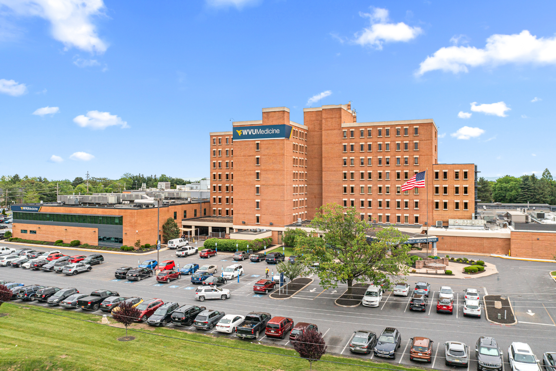Aerial view of WVU Medicine Berkeley Medical Center with parking lot and American flag on a sunny day.