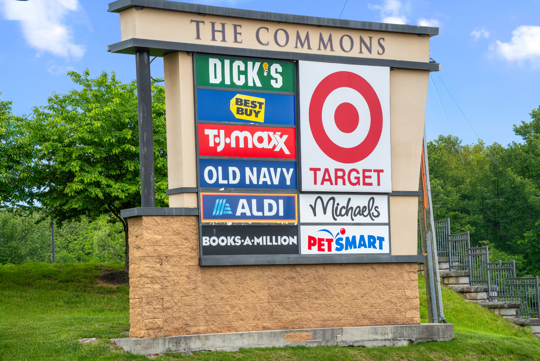 Shopping center sign featuring stores like Dick\'s Sporting Goods, Best Buy, Target, PetSmart, and more against a green, outdoor setting.