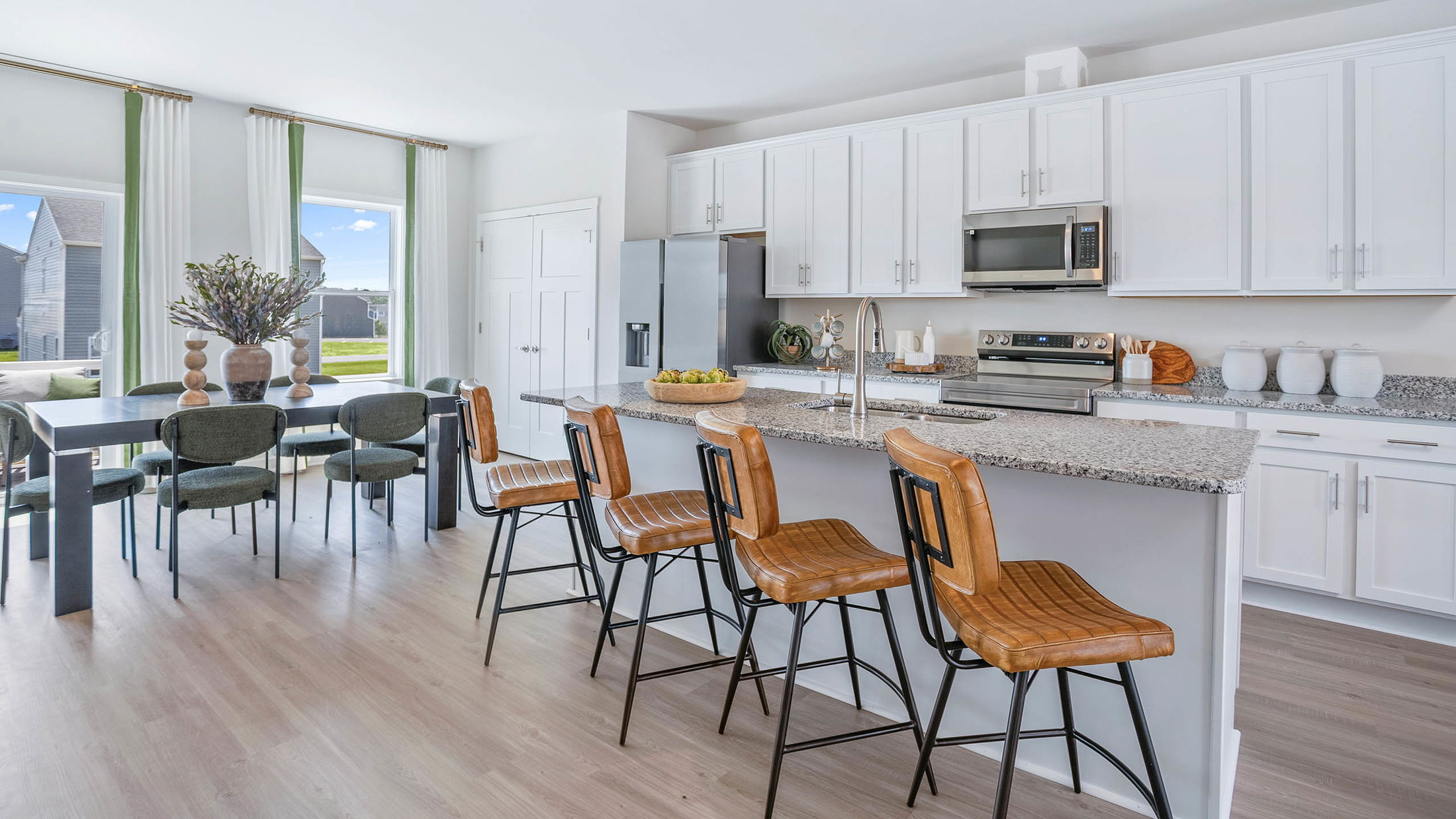 Modern kitchen with white cabinets, granite countertops, and leather bar stools, adjacent to a dining area with large windows and stylish decor.