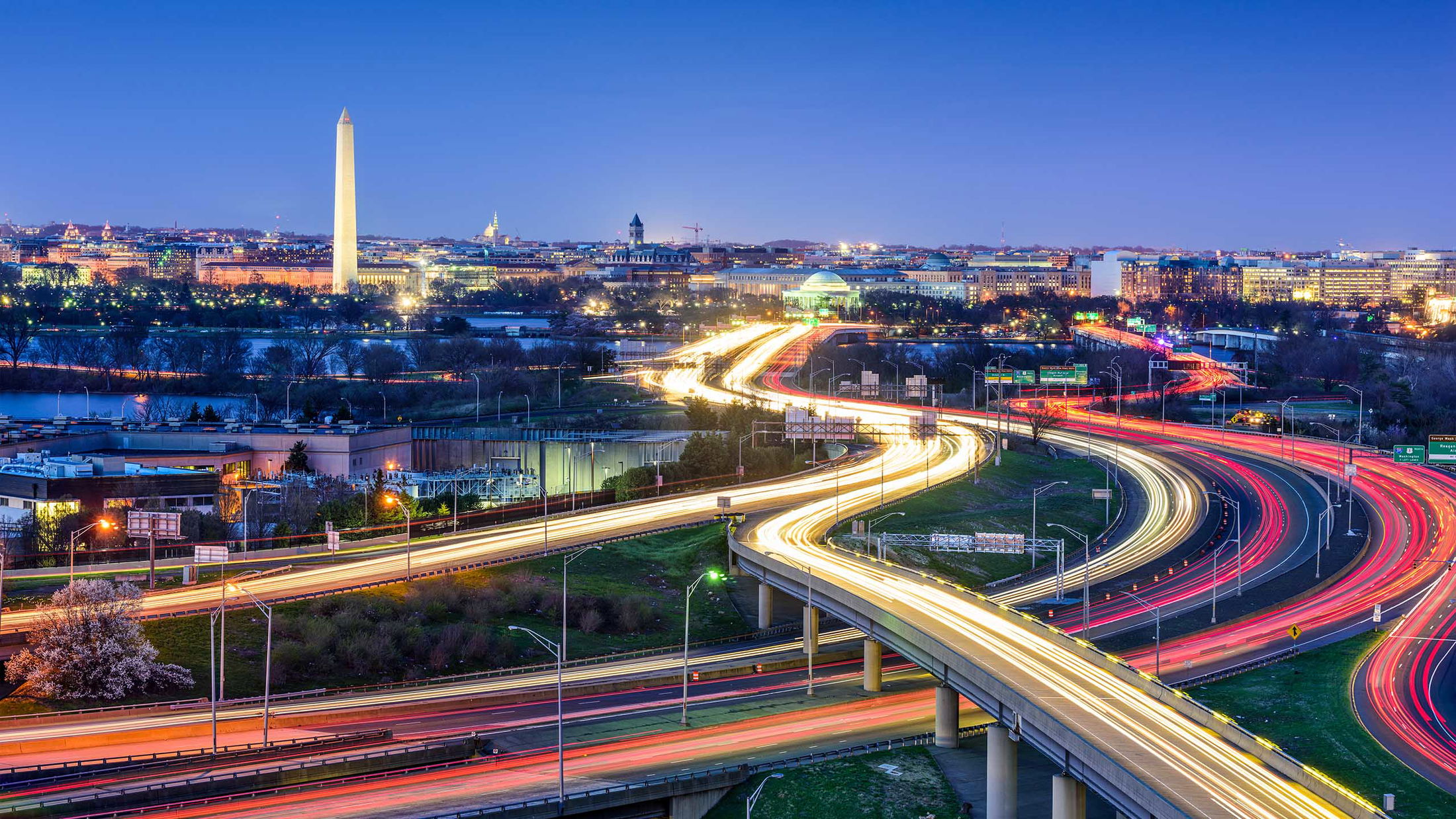 Night view of Washington, D.C.'s illuminated highways leading towards the Washington Monument against a vibrant city skyline.