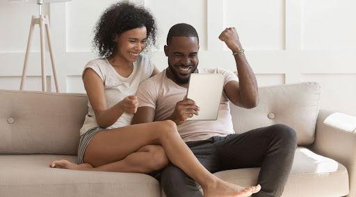 A happy couple sitting on a couch celebrating while looking at a tablet.