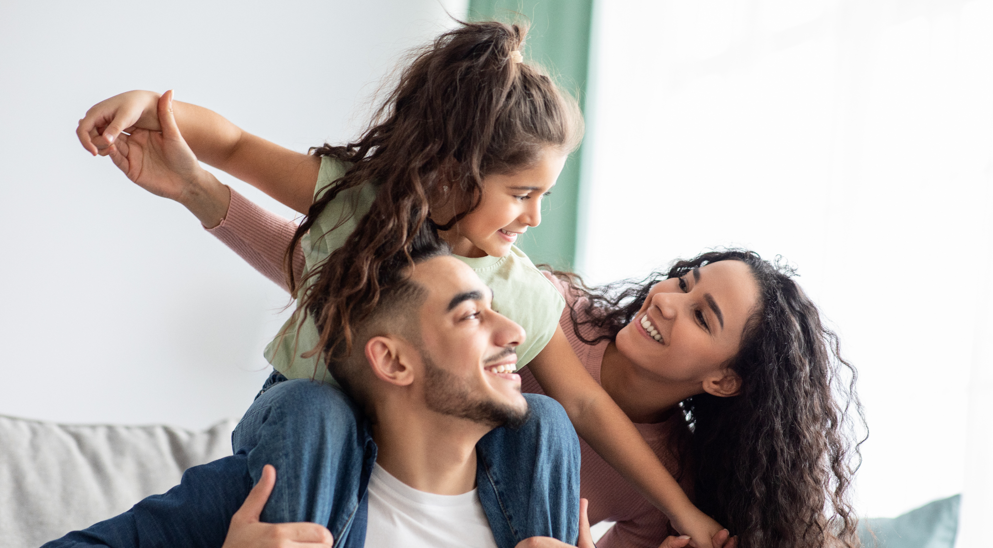 Happy family playing together in a bright living room, with a young girl playfully sitting on a man\\\'s shoulders.
