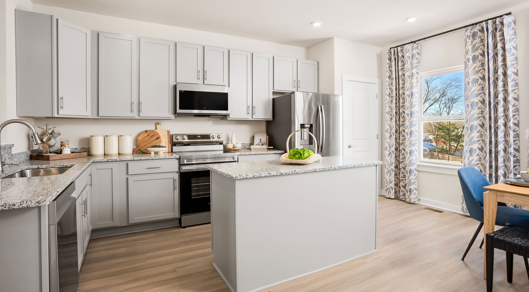 Modern kitchen interior with light gray cabinets, stainless steel appliances, a central island, and a view of blue patterned curtains.
