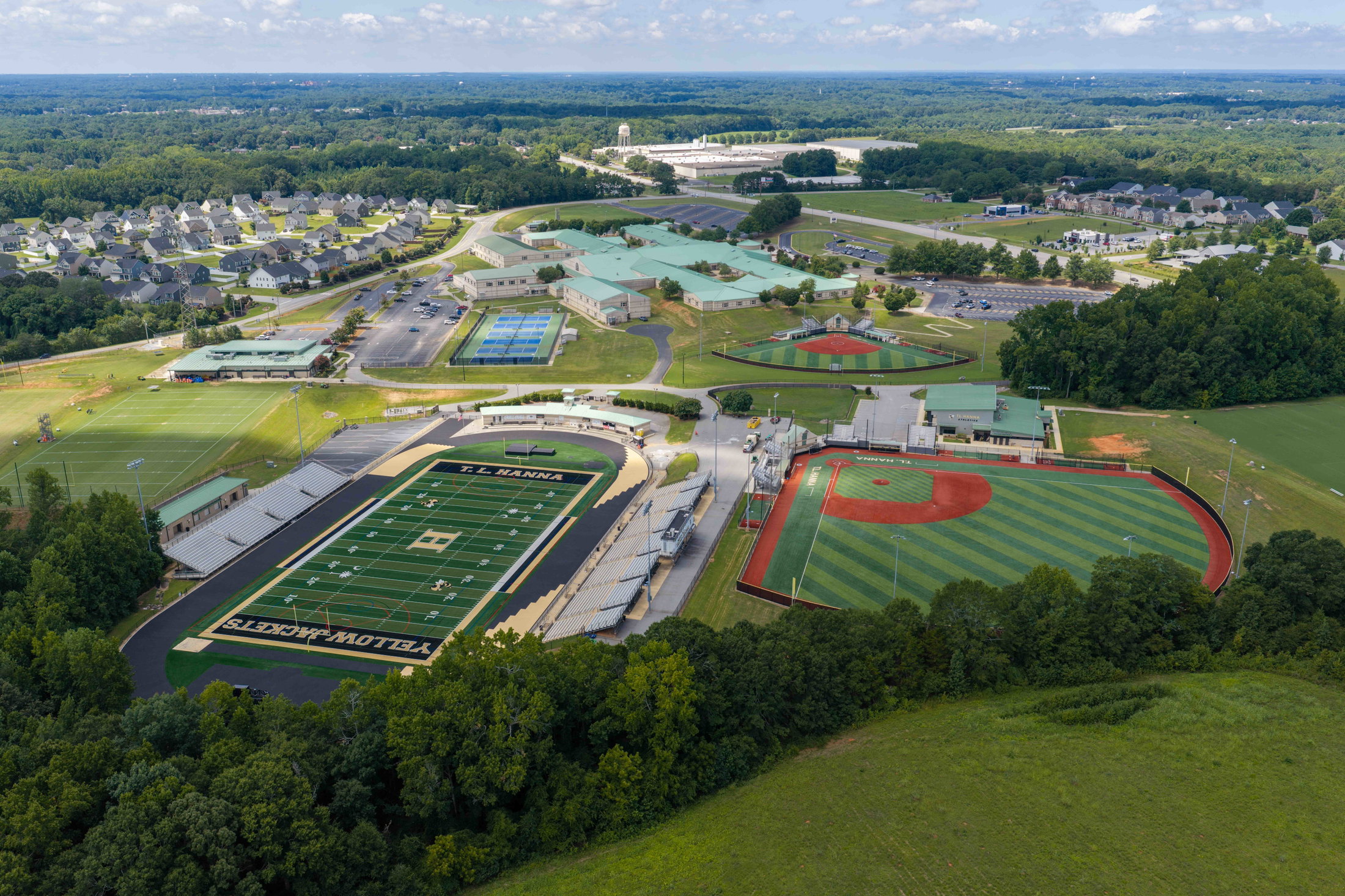 Aerial view of a high school with a football field and baseball diamond surrounded by green fields and residential housing.