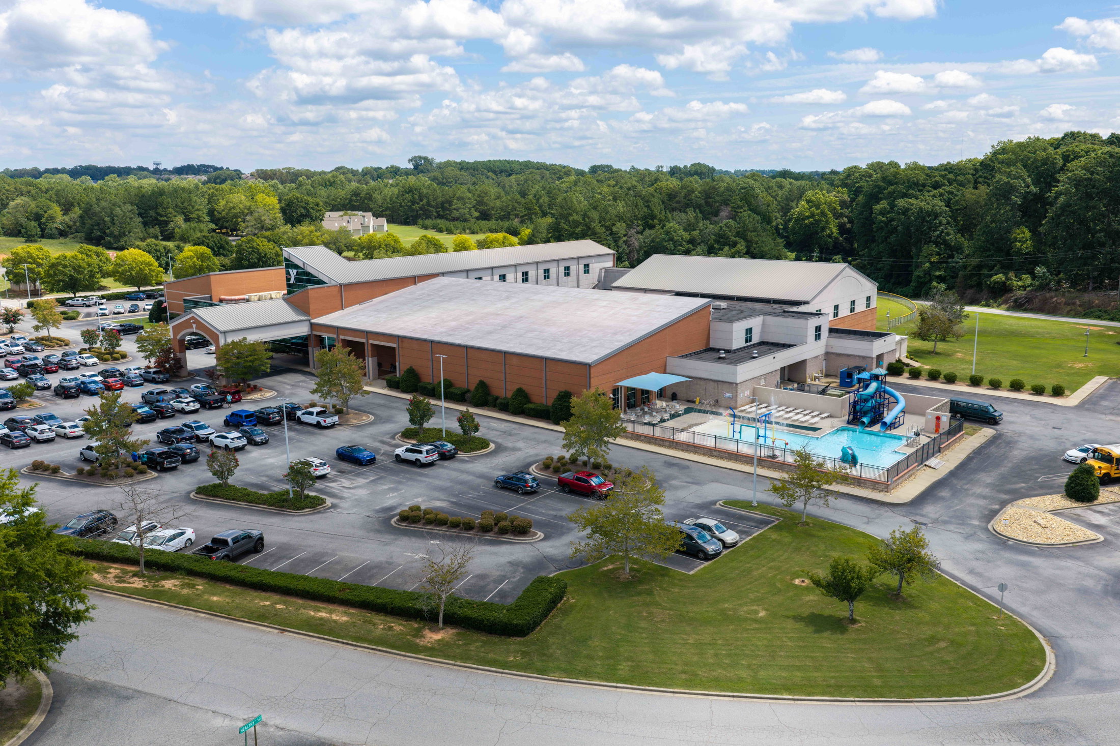 Aerial view of a recreational facility with a pool and parking lot surrounded by trees.