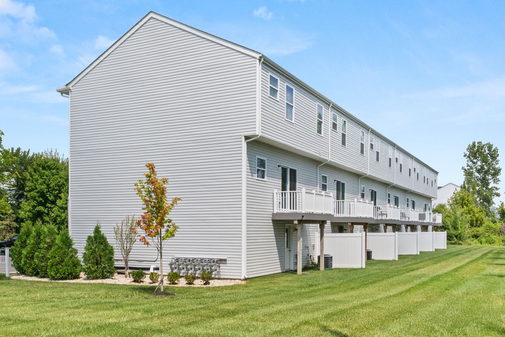Row of modern townhouses with balconies, surrounded by neatly landscaped greenery against a clear blue sky.