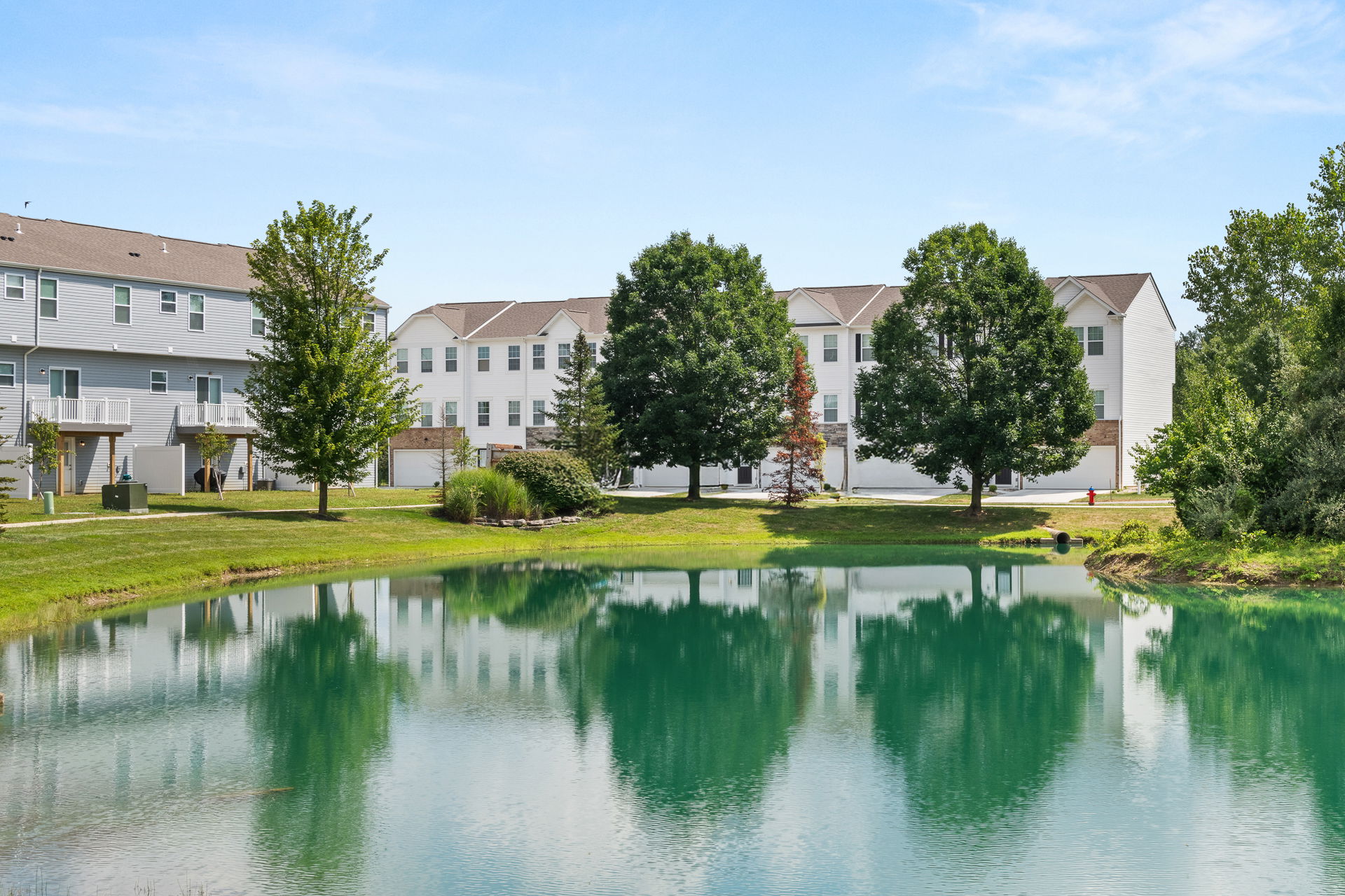 Townhouses and trees reflected in a peaceful pond under a clear blue sky.