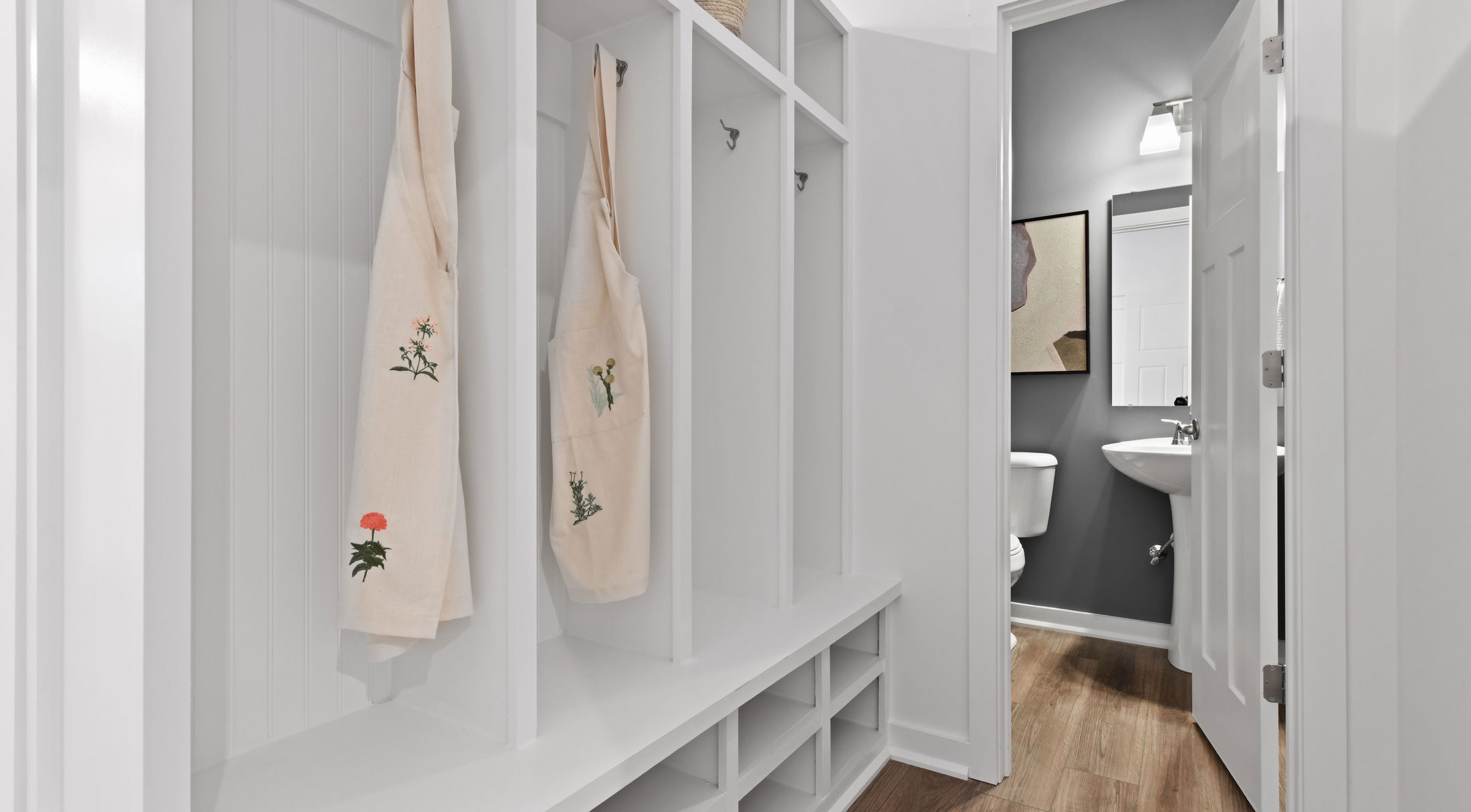 Modern mudroom with white lockers and hooks, featuring floral aprons, beside a sleek bathroom with gray walls and wooden flooring.