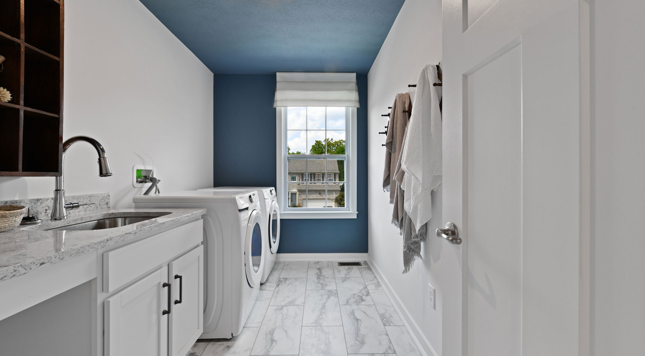 Modern laundry room with white cabinetry, a sleek countertop with a stainless steel sink, front-loading washer and dryer, and a bright window.