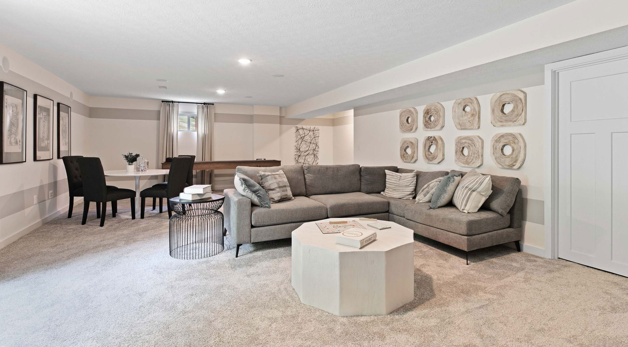 Modern basement living room with a gray sectional sofa, abstract wall art, and a white octagonal coffee table on a plush carpet.