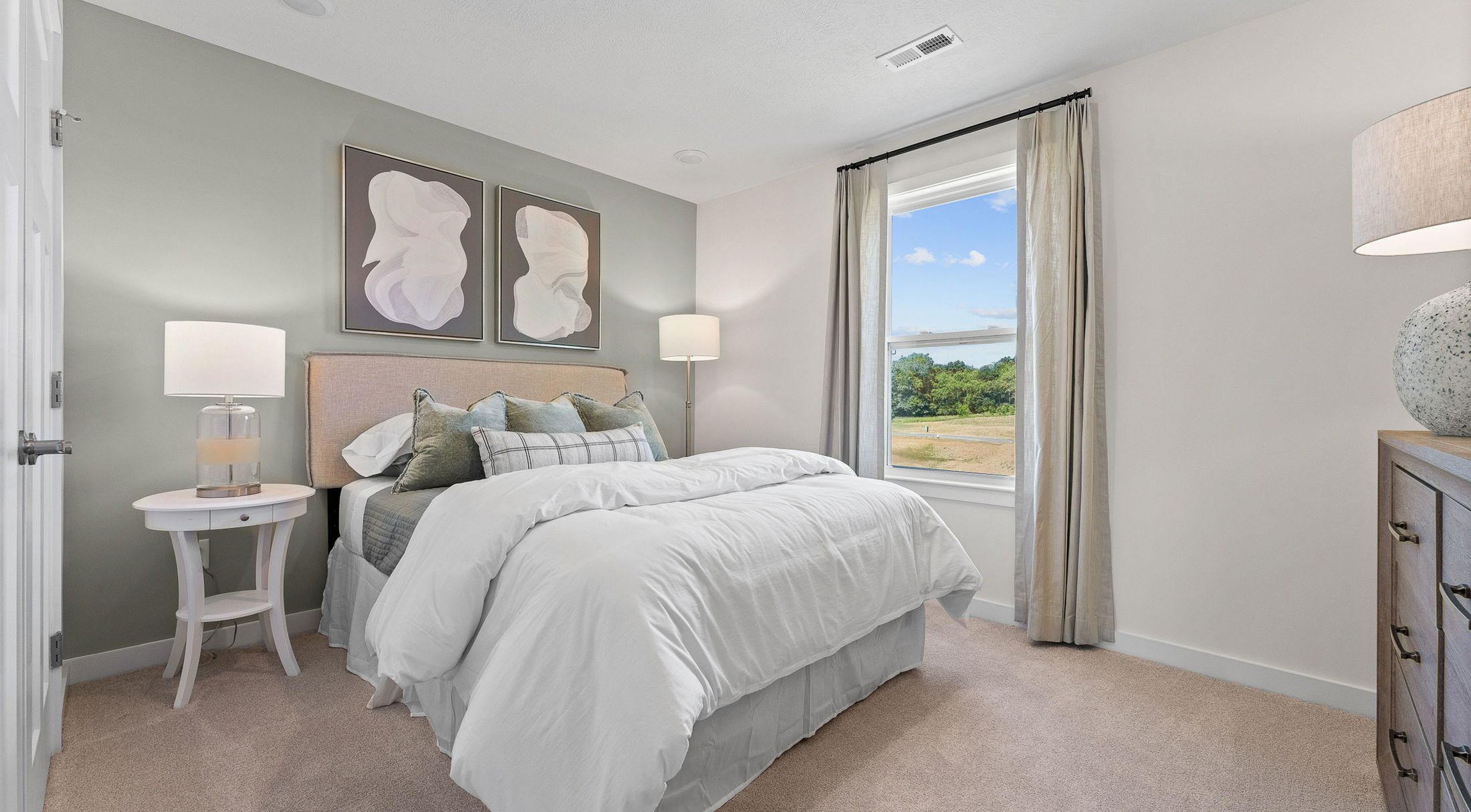 Modern bedroom interior featuring a cozy bed with white linens, accent pillows, bedside tables with lamps, abstract wall art, and a large window with scenic views.