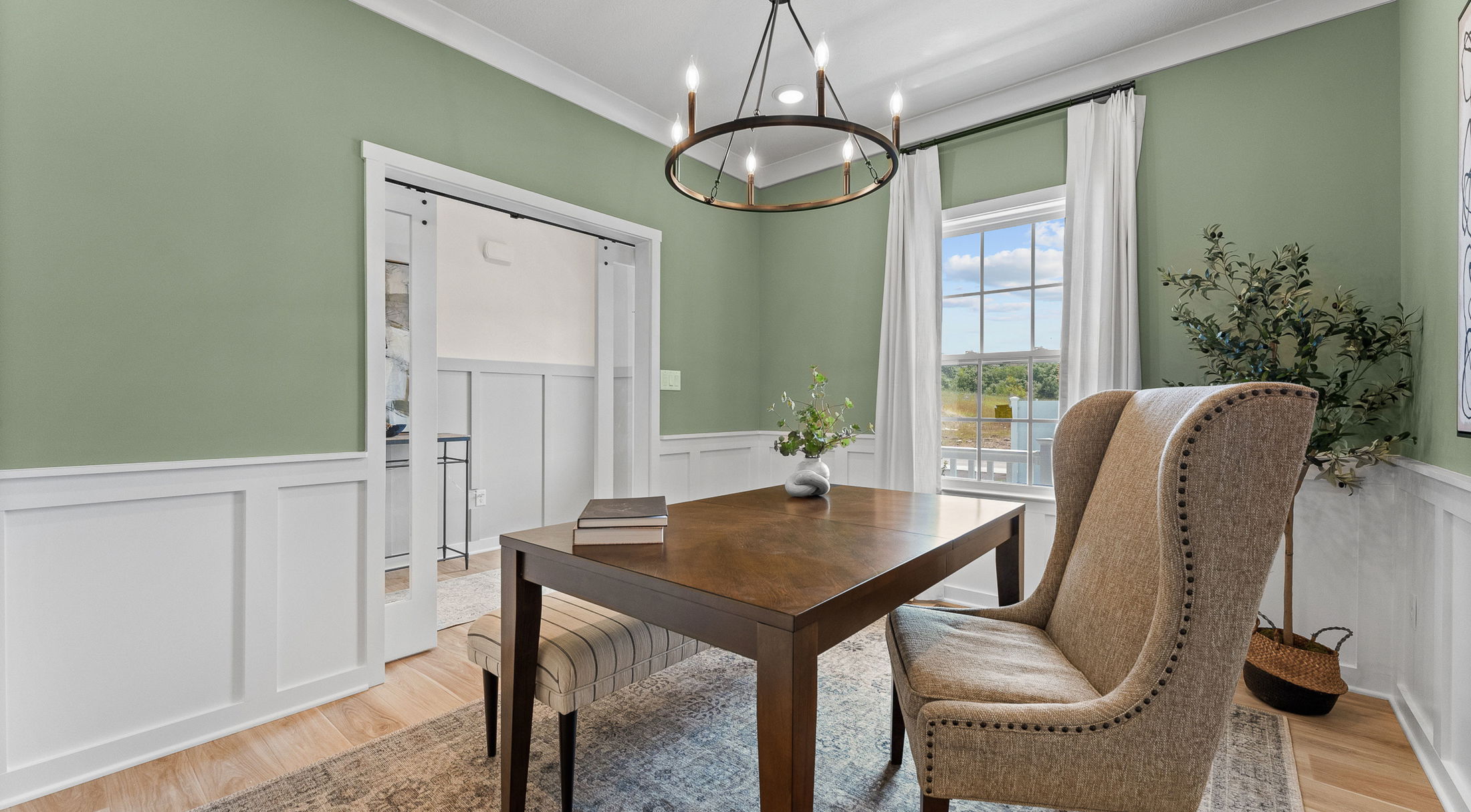 Elegant home office with green walls, wooden desk, upholstered chairs, and modern chandelier.