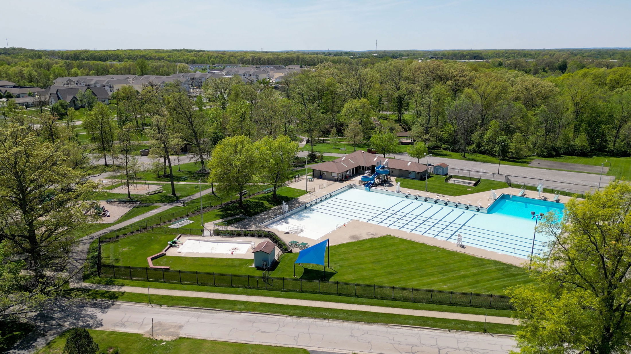 Aerial view of a community park with a large outdoor swimming pool surrounded by green trees and houses.