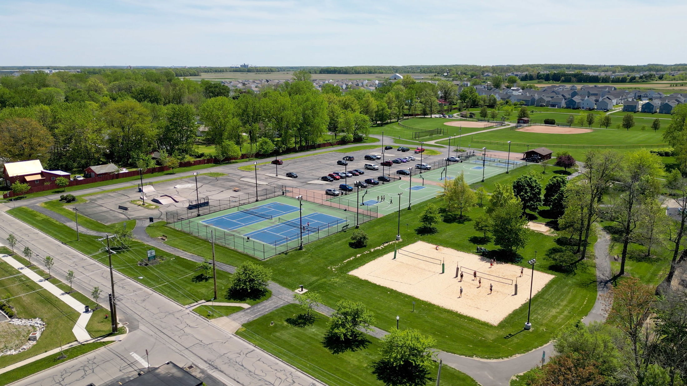 Aerial view of a recreational park featuring tennis courts, a skate park, sand volleyball courts, and a baseball field surrounded by green landscape.