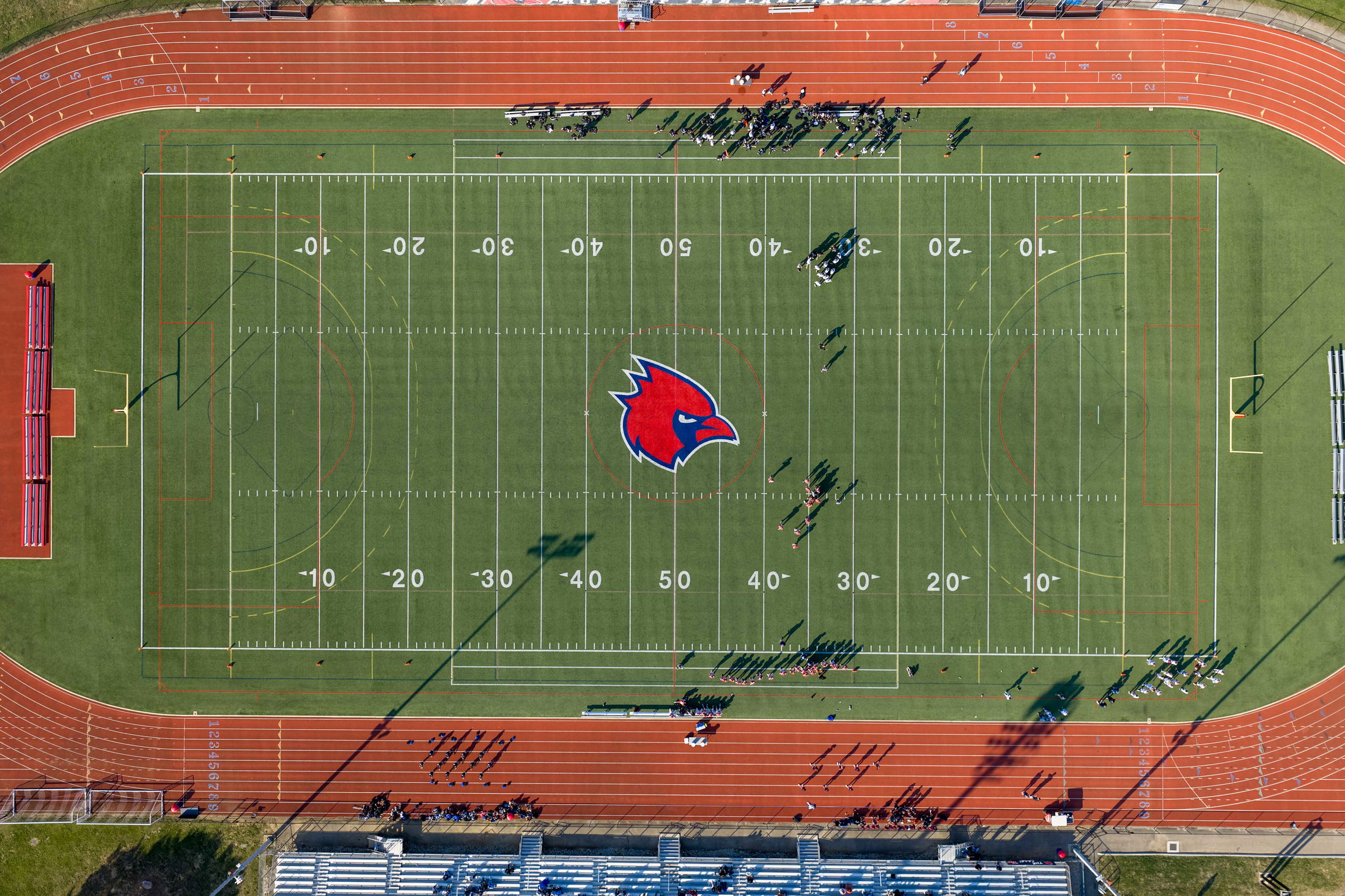 Aerial view of a football field with a red cardinal logo at the center, surrounded by a running track and scattered people.