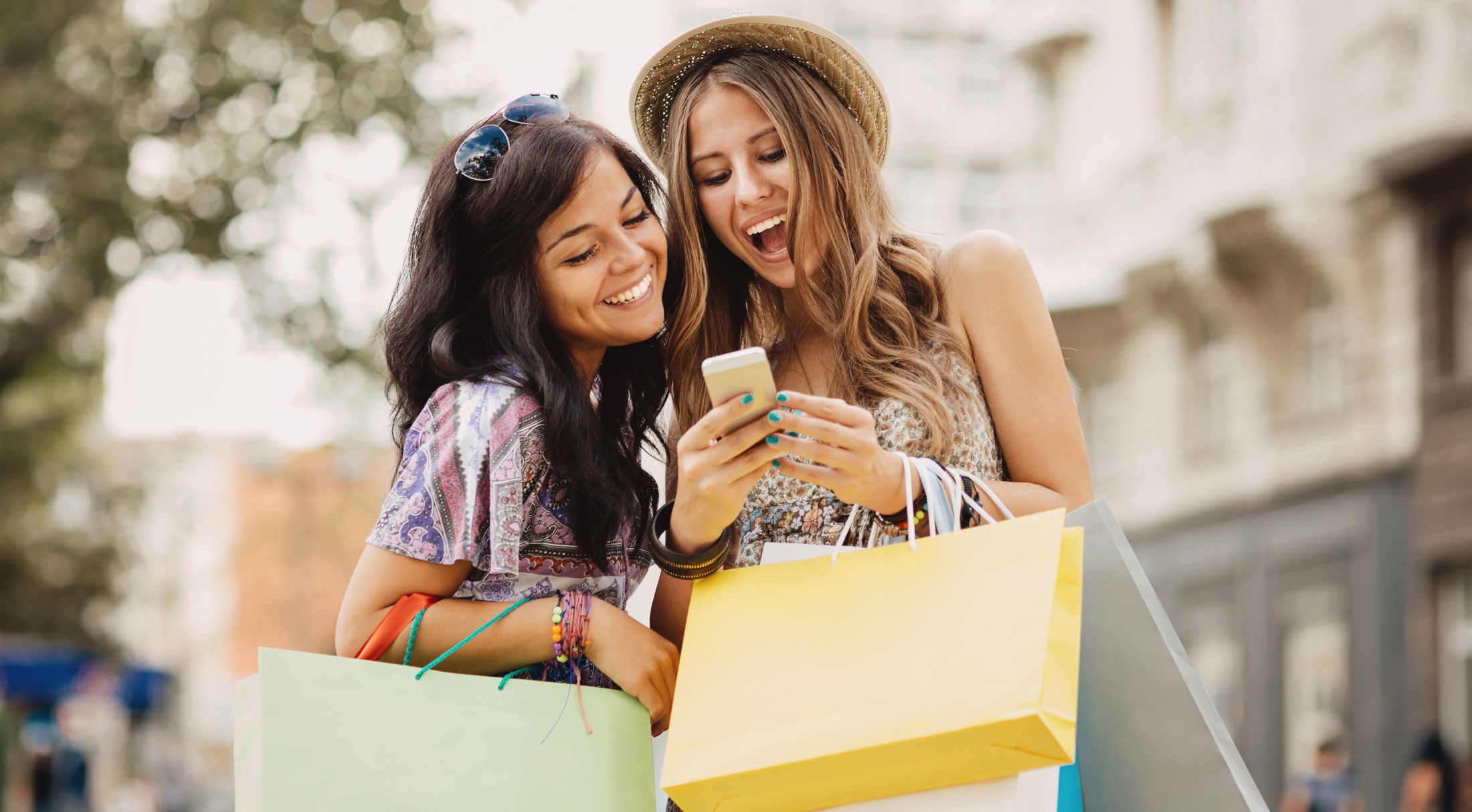 Two happy young women with shopping bags smiling and looking at a smartphone outdoors.