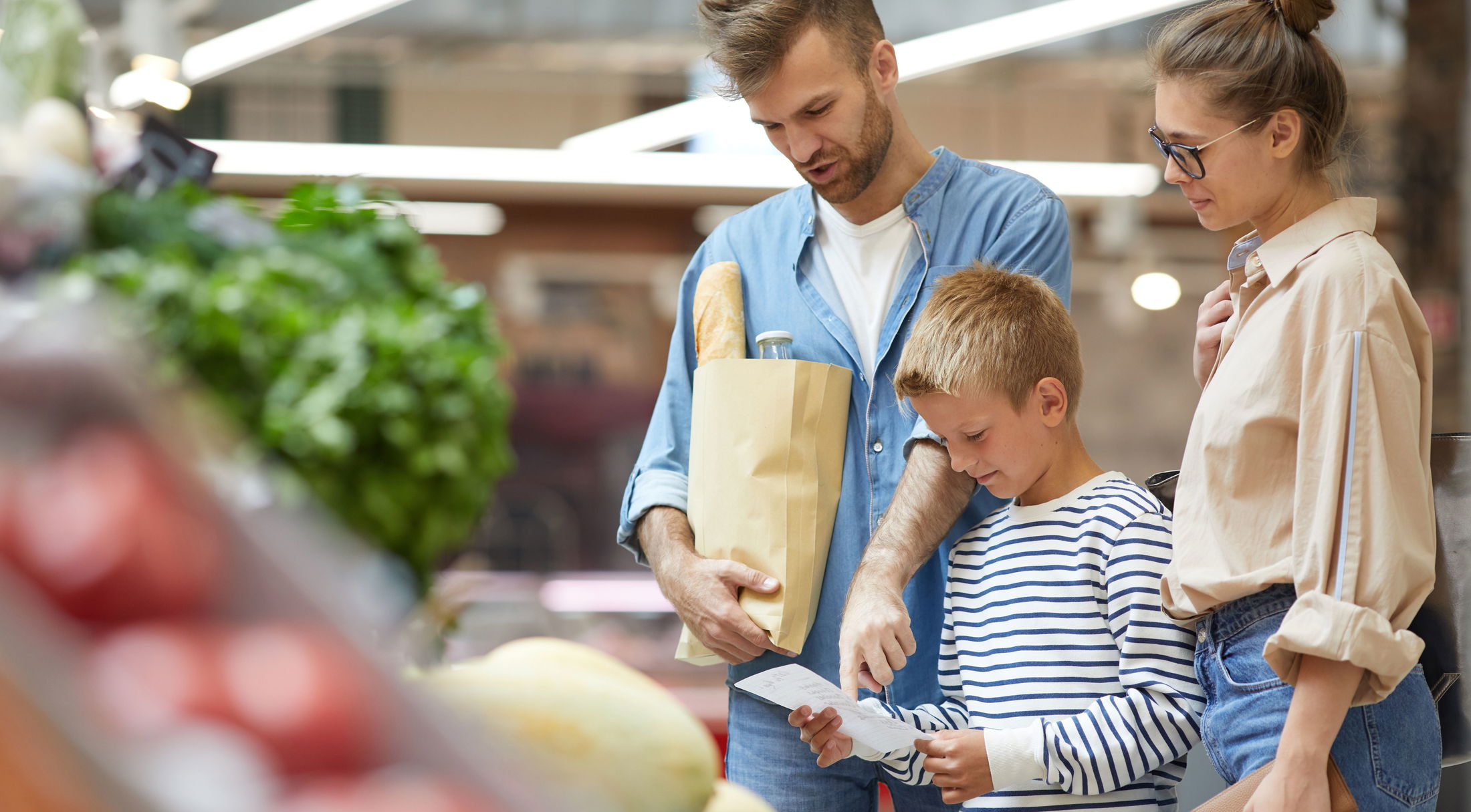 Family grocery shopping at a supermarket, with a man holding a paper bag filled with groceries while a woman and boy look at a receipt.