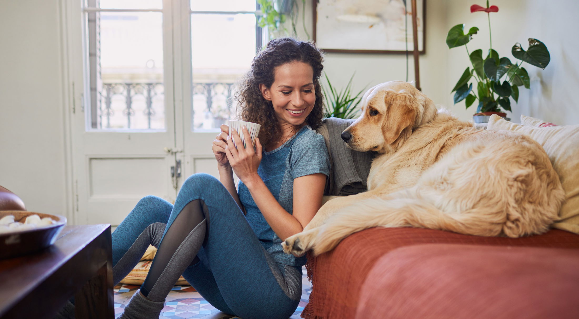 A woman in a blue outfit enjoys a cozy moment with her golden retriever on the couch, holding a cup of coffee in a sunlit living room.