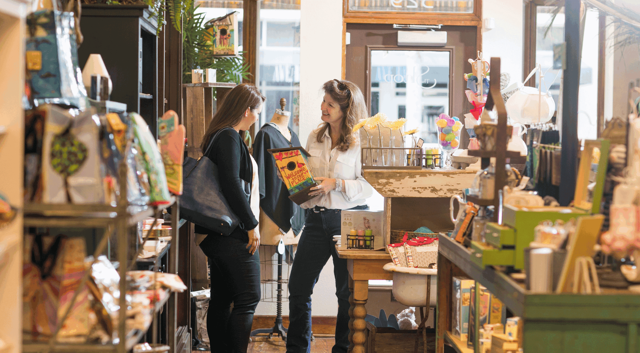 Two women engaging in a friendly conversation while browsing a cozy boutique shop filled with colorful and eclectic items.