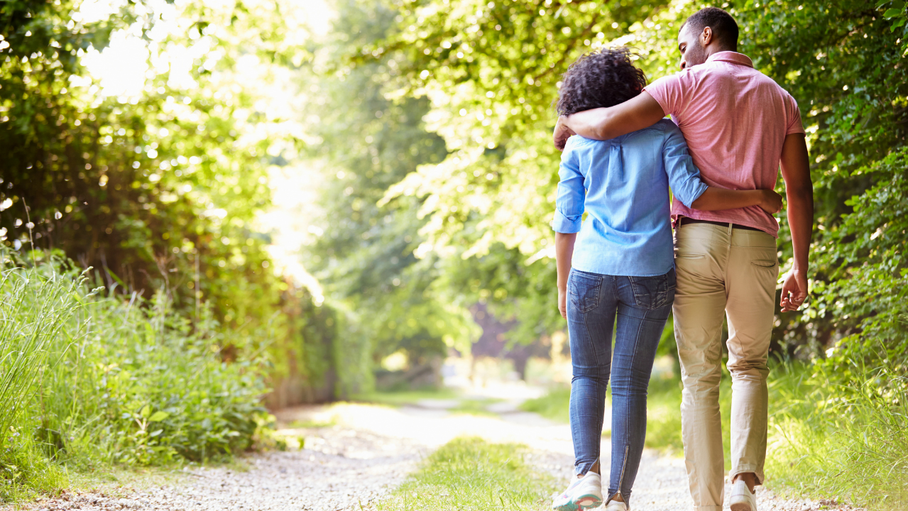 A couple walking arm in arm down a sunlit forest path with lush green trees on a summer day.