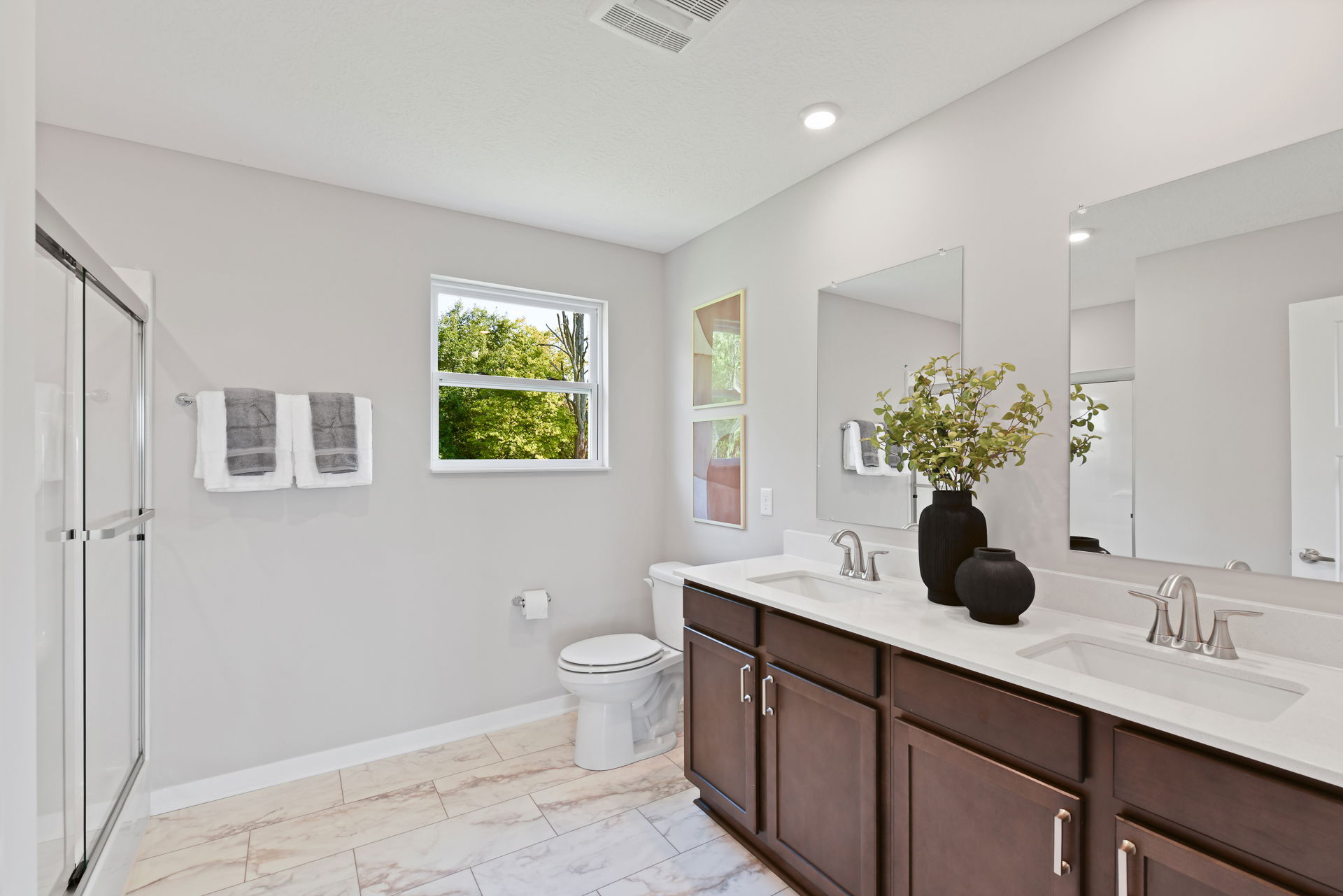 Modern bathroom with dual sinks, large mirrors, a toilet, and a glass-enclosed shower, featuring sleek fixtures and natural light from a window.