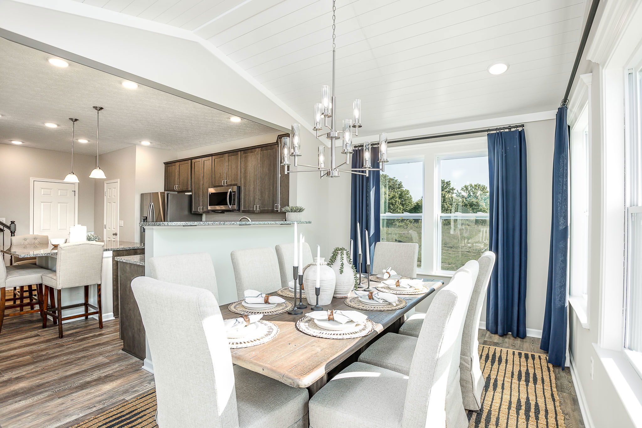 Modern open-concept dining and kitchen area with light-colored furnishings and natural light from large windows.
