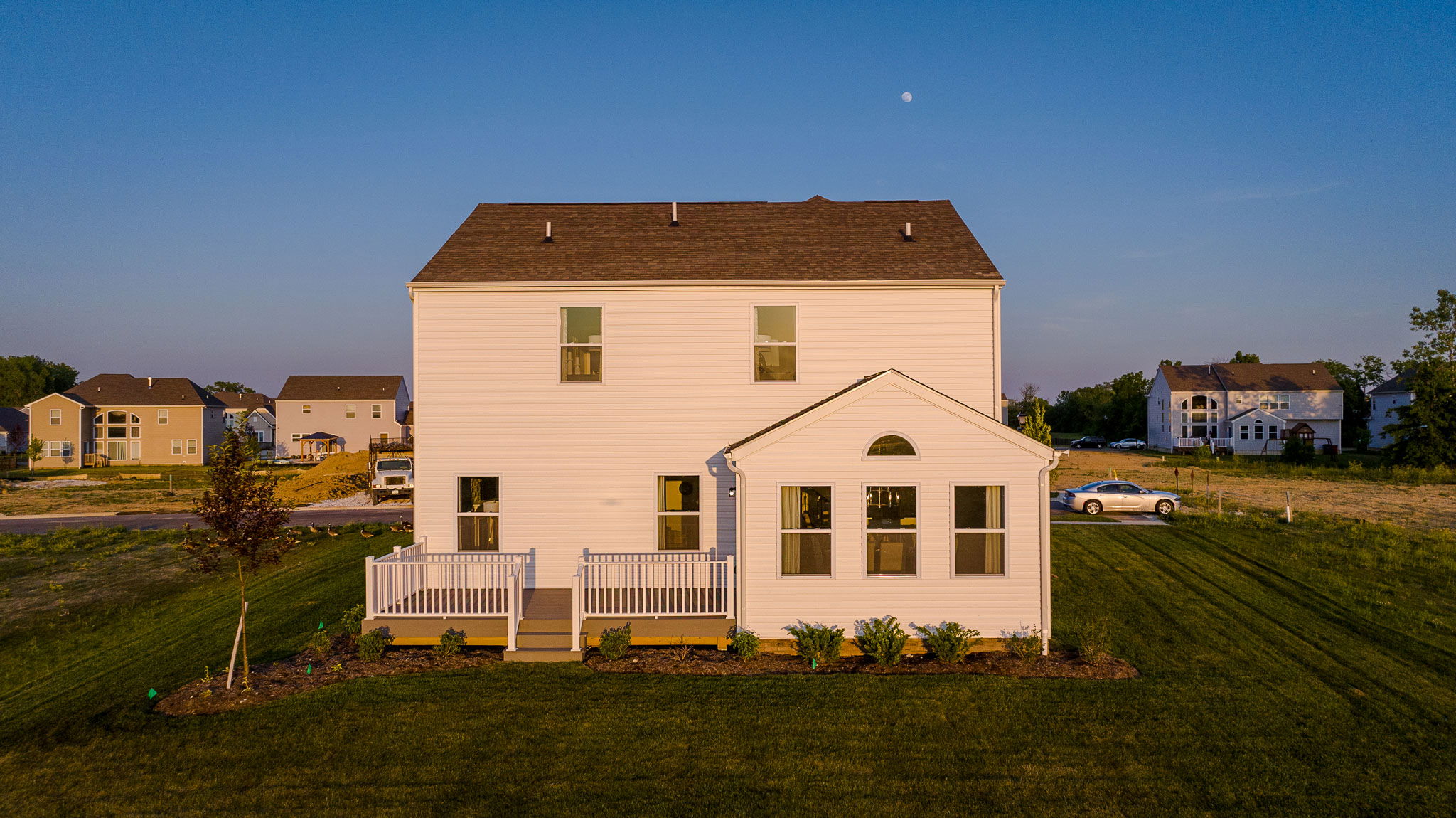 Back view of a suburban two-story white house with a manicured lawn and neighboring homes at sunset.