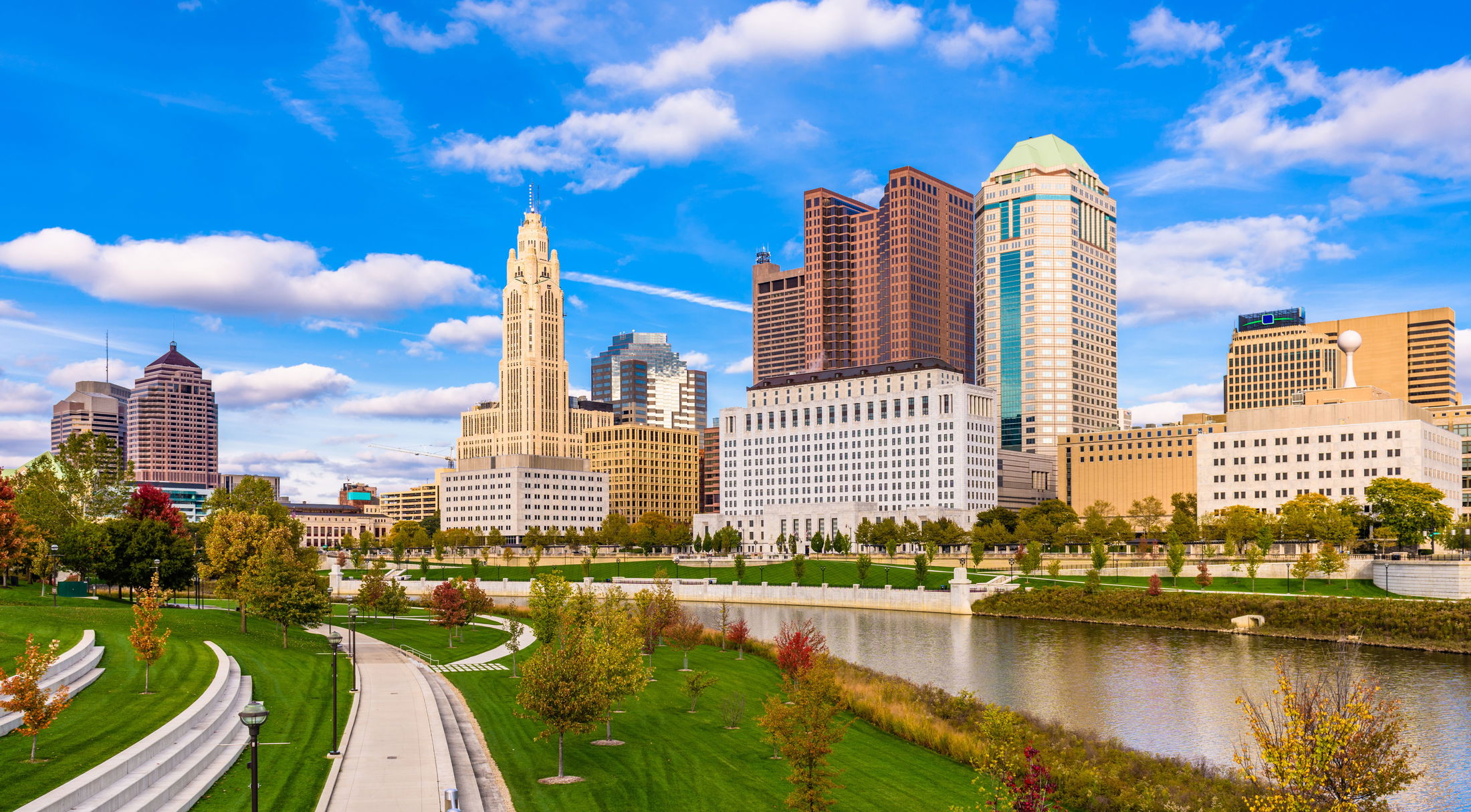 Skyline of downtown Columbus, Ohio, featuring modern skyscrapers and a scenic riverfront park under a vibrant blue sky.