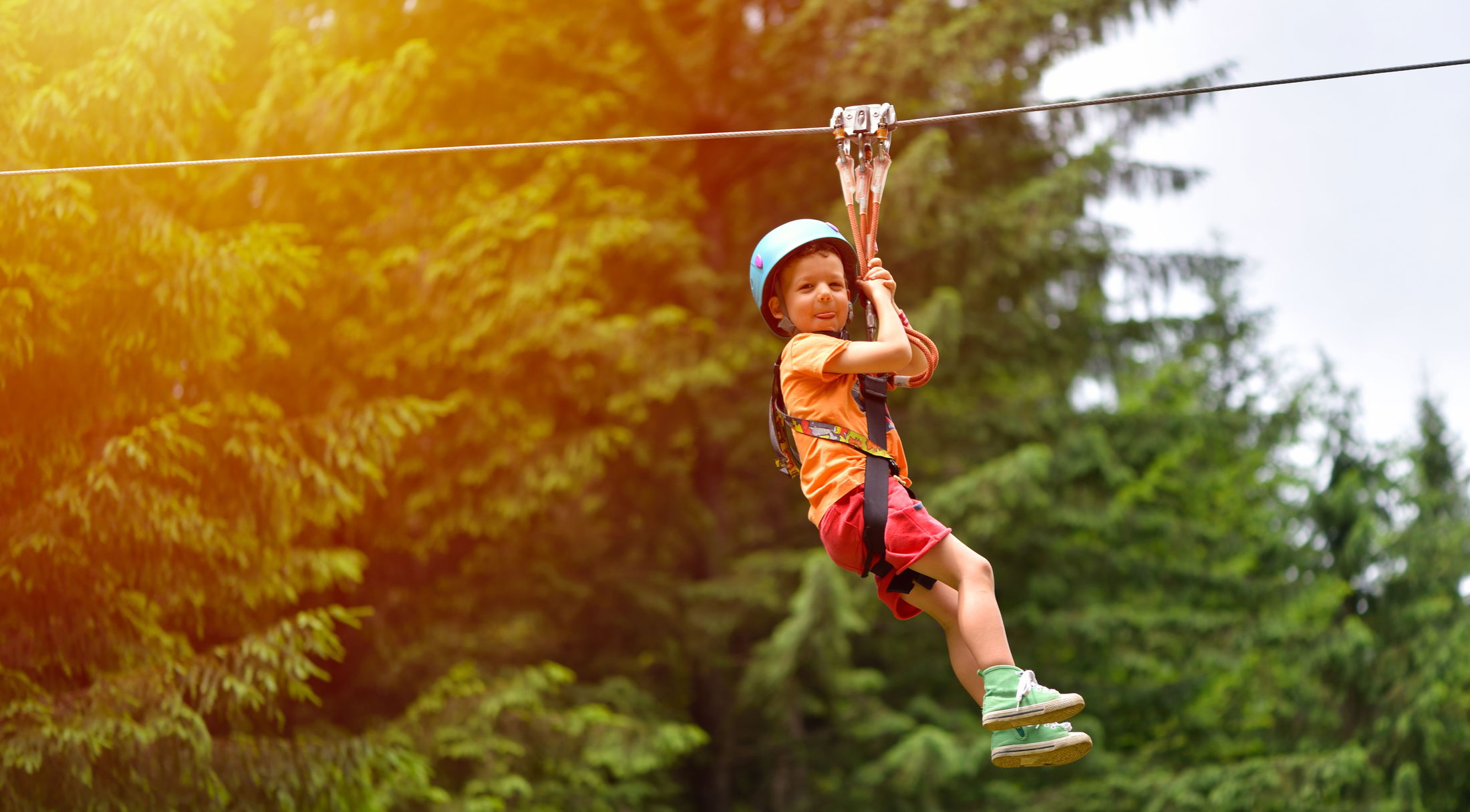 A young child zip-lining through a lush forest wearing a blue helmet and safety harness.