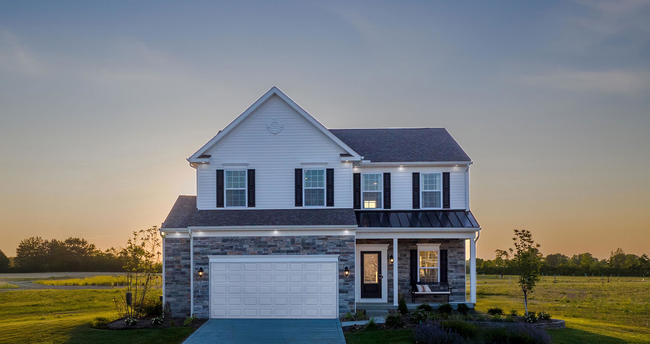 Two-story suburban house with a large garage, set against a sunset sky in a landscaped field.