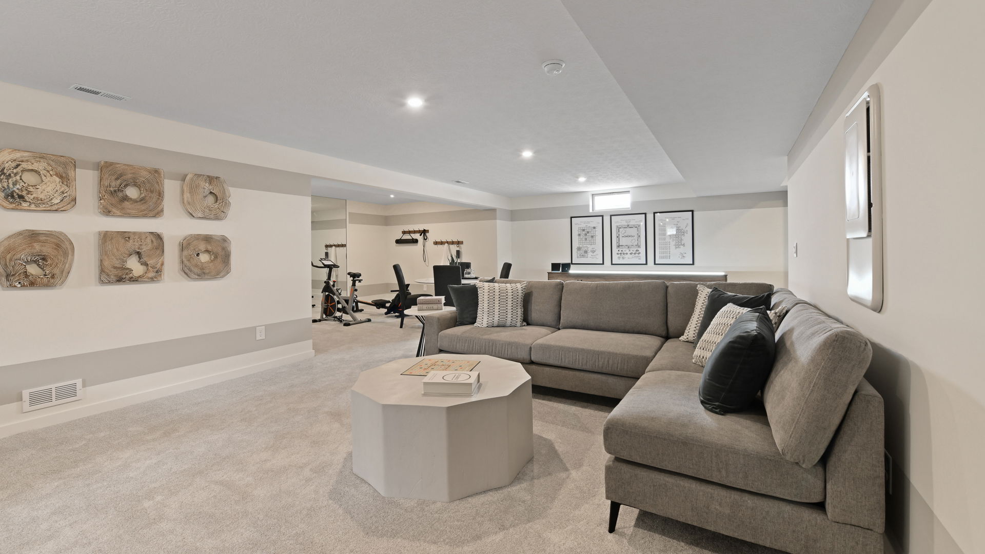 Spacious modern basement with a plush gray sectional sofa, gym equipment, and minimalist wall art.