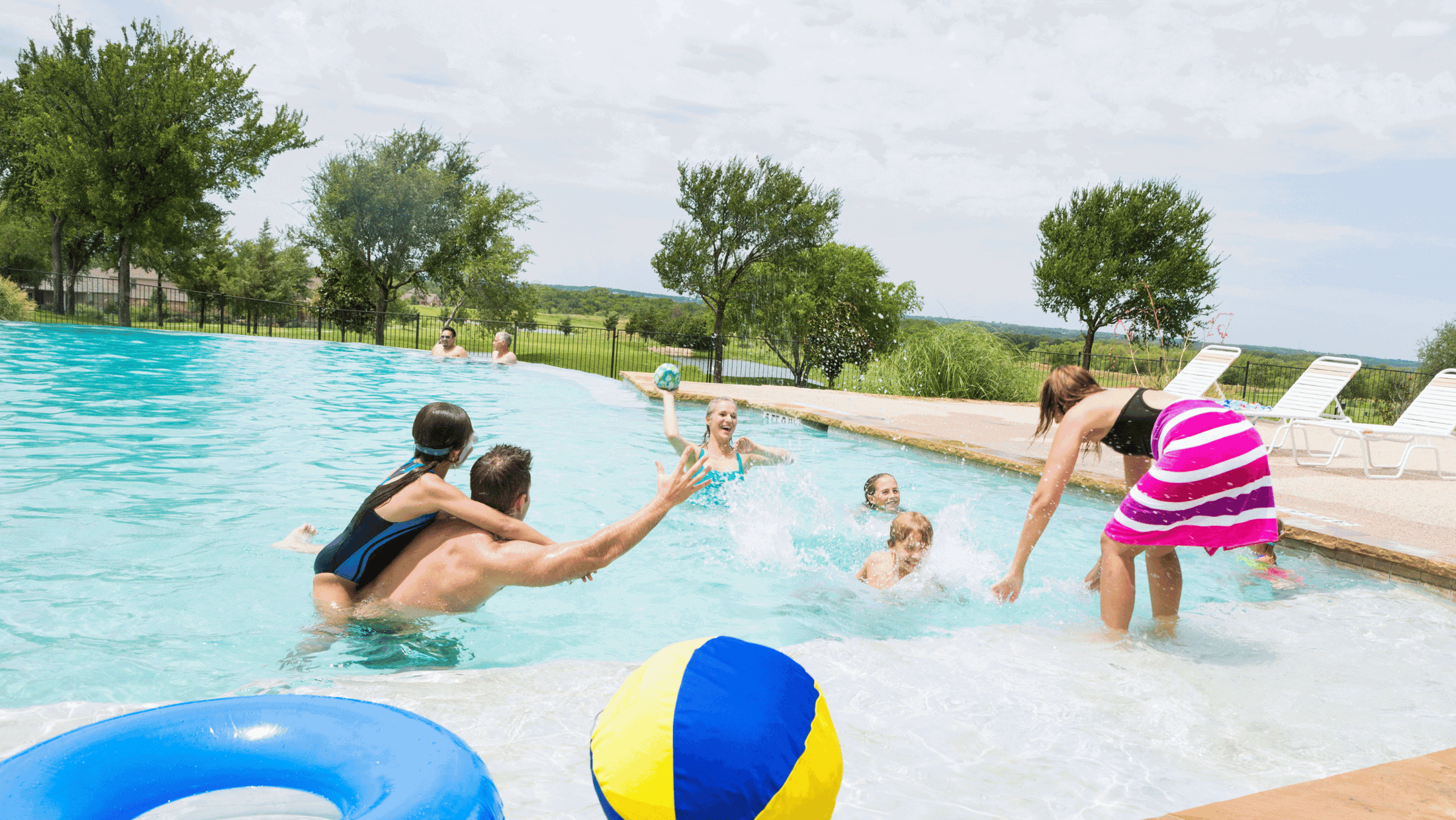 A group of people enjoying a lively game of water volleyball in a beautiful outdoor pool setting on a sunny day, surrounded by greenery and poolside chairs.