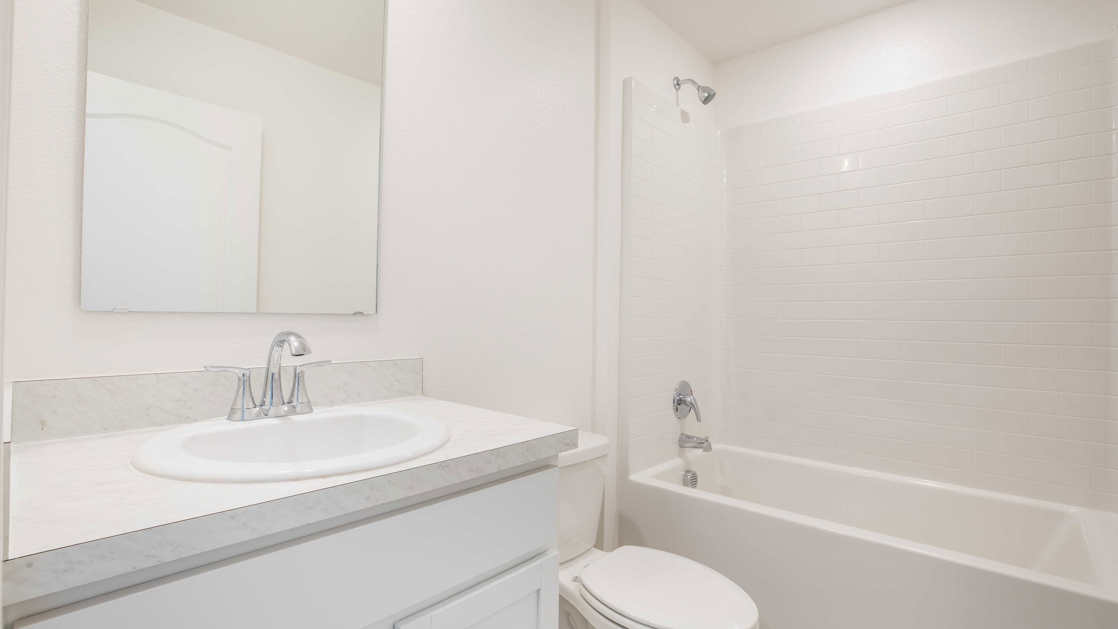 A modern, bright white bathroom featuring a sleek vanity with a marble countertop, a wall mirror, a toilet, and a classic bathtub with a tile surround.