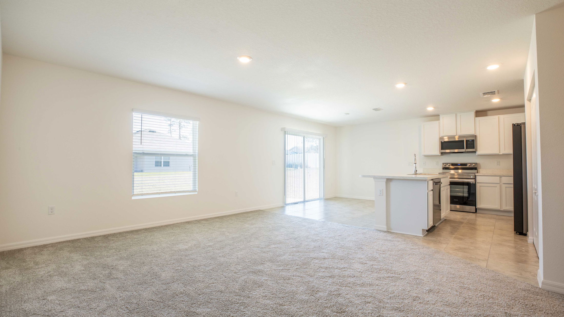 Modern open-concept living room and kitchen with light carpeting, white cabinets, and stainless steel appliances.