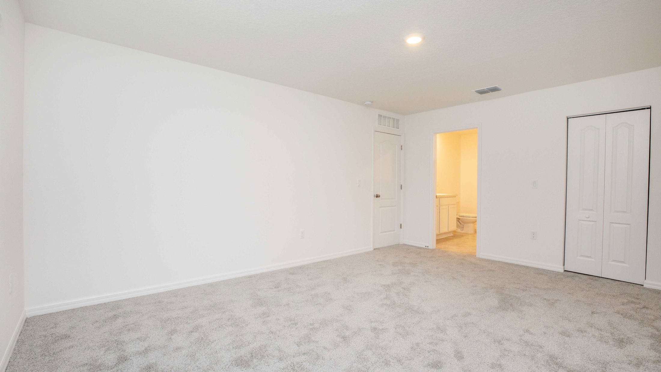 Spacious empty room with beige carpeting, white walls, and an open door leading to a bathroom.