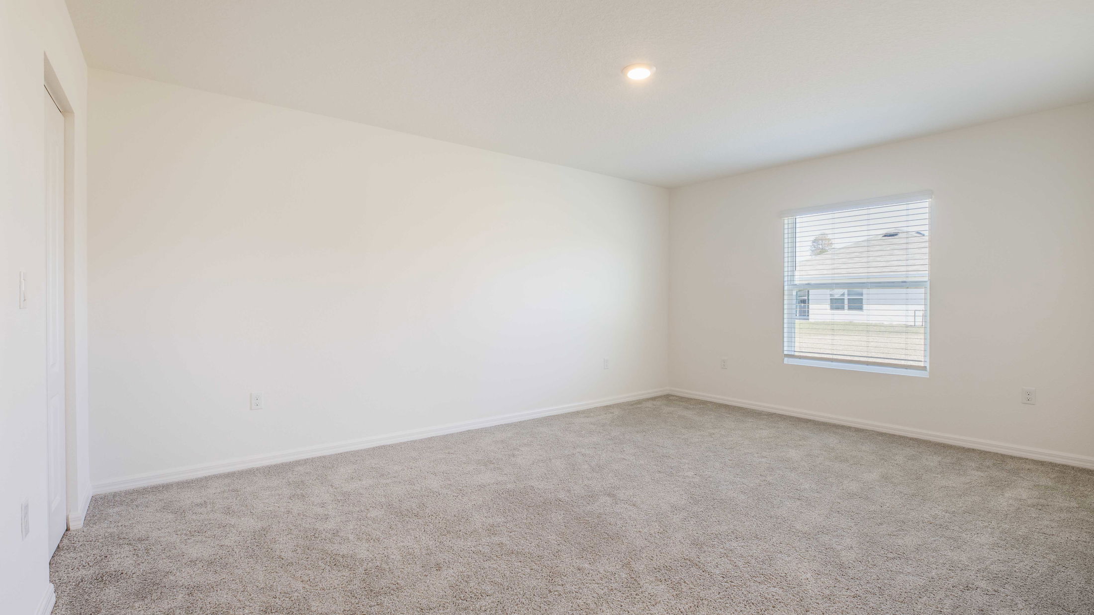 Empty room with beige carpet, white walls, a window with blinds, and a ceiling light, showcasing interior design potential.
