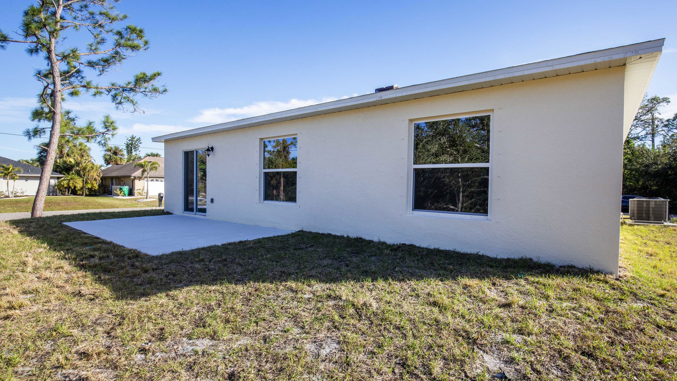 Backyard view of a beige single-story house with large windows, a concrete patio, and a grassy yard under a clear blue sky.