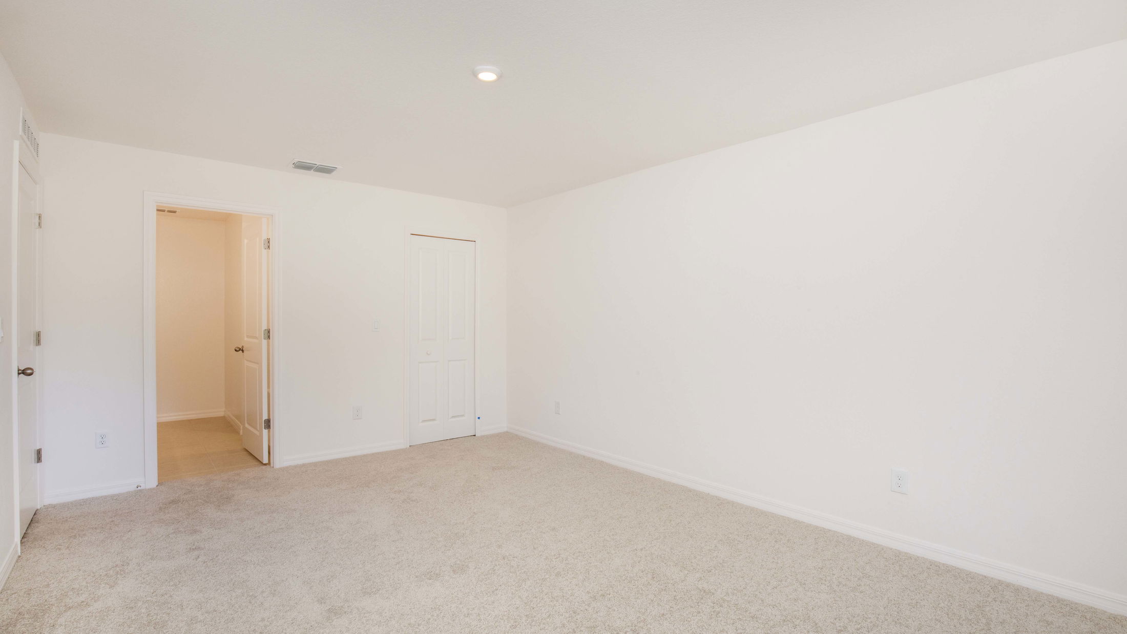 Empty, spacious bedroom with beige carpet, white walls, and an open door leading to another room.