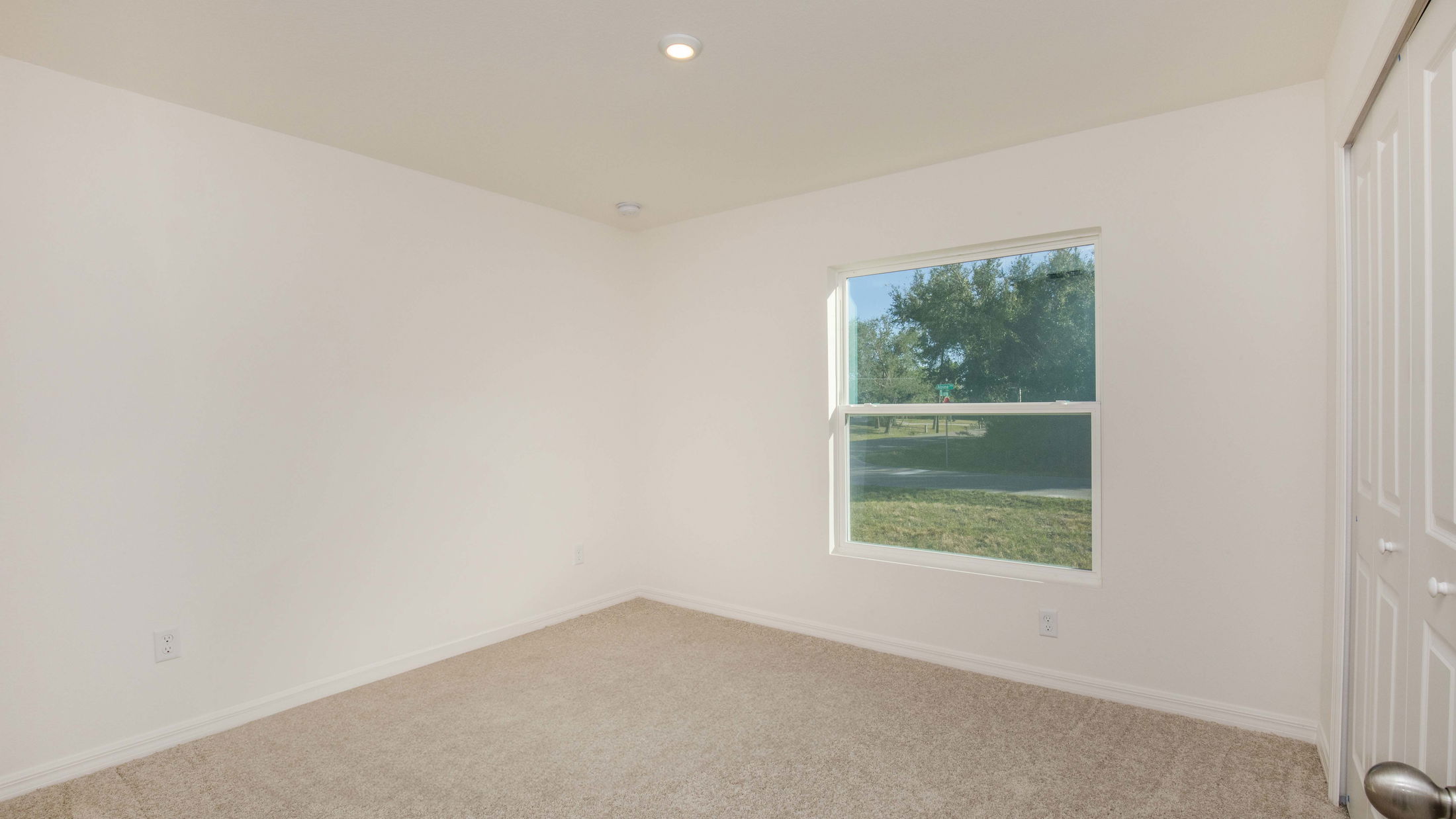 A bright, empty bedroom with beige carpet, white walls, and a large window overlooking a green lawn.