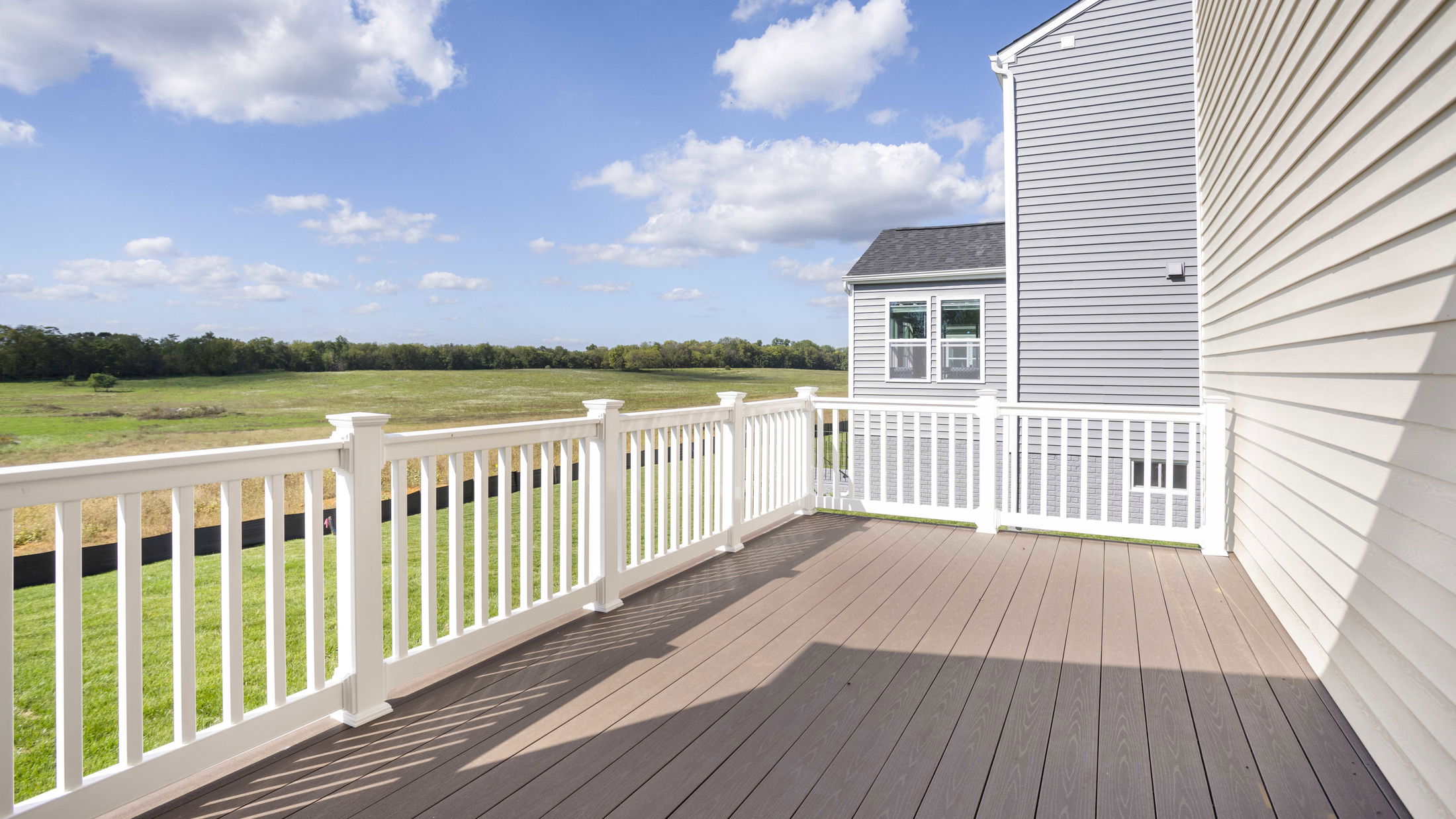 Spacious outdoor deck with white railing on a sunny day, overlooking a green field and blue sky.