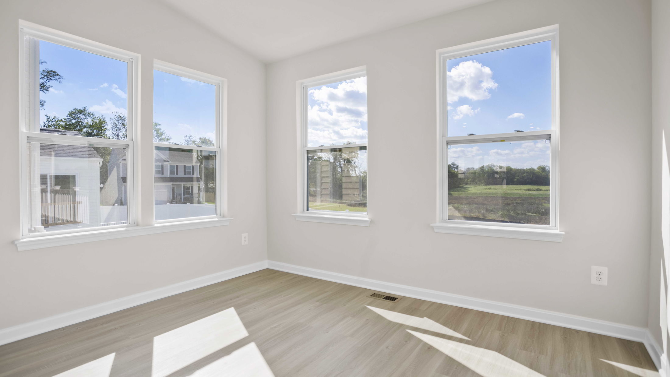 Bright, empty room with three large windows and light wood flooring showcasing a clear view of a sunny exterior landscape.