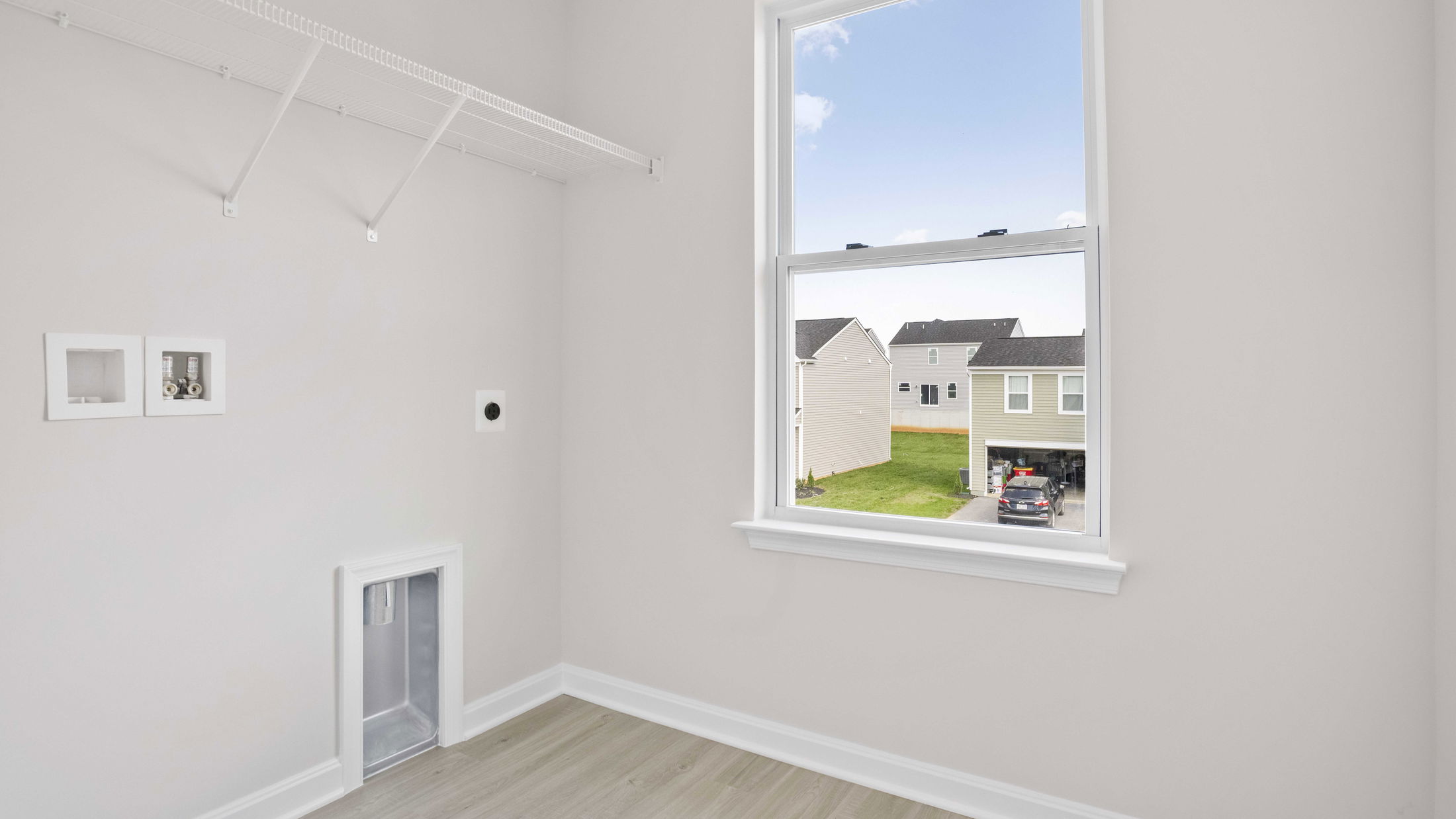 Empty laundry room with beige walls, a window overlooking suburban houses, and wall-mounted hookups for washer and dryer installation.