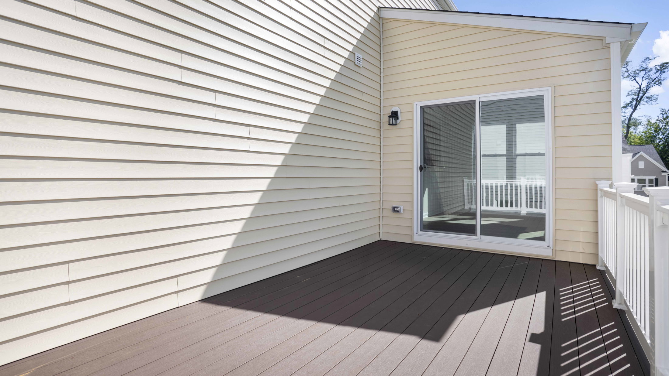 Sunlit beige vinyl siding and wooden deck with sliding glass door on a residential balcony.