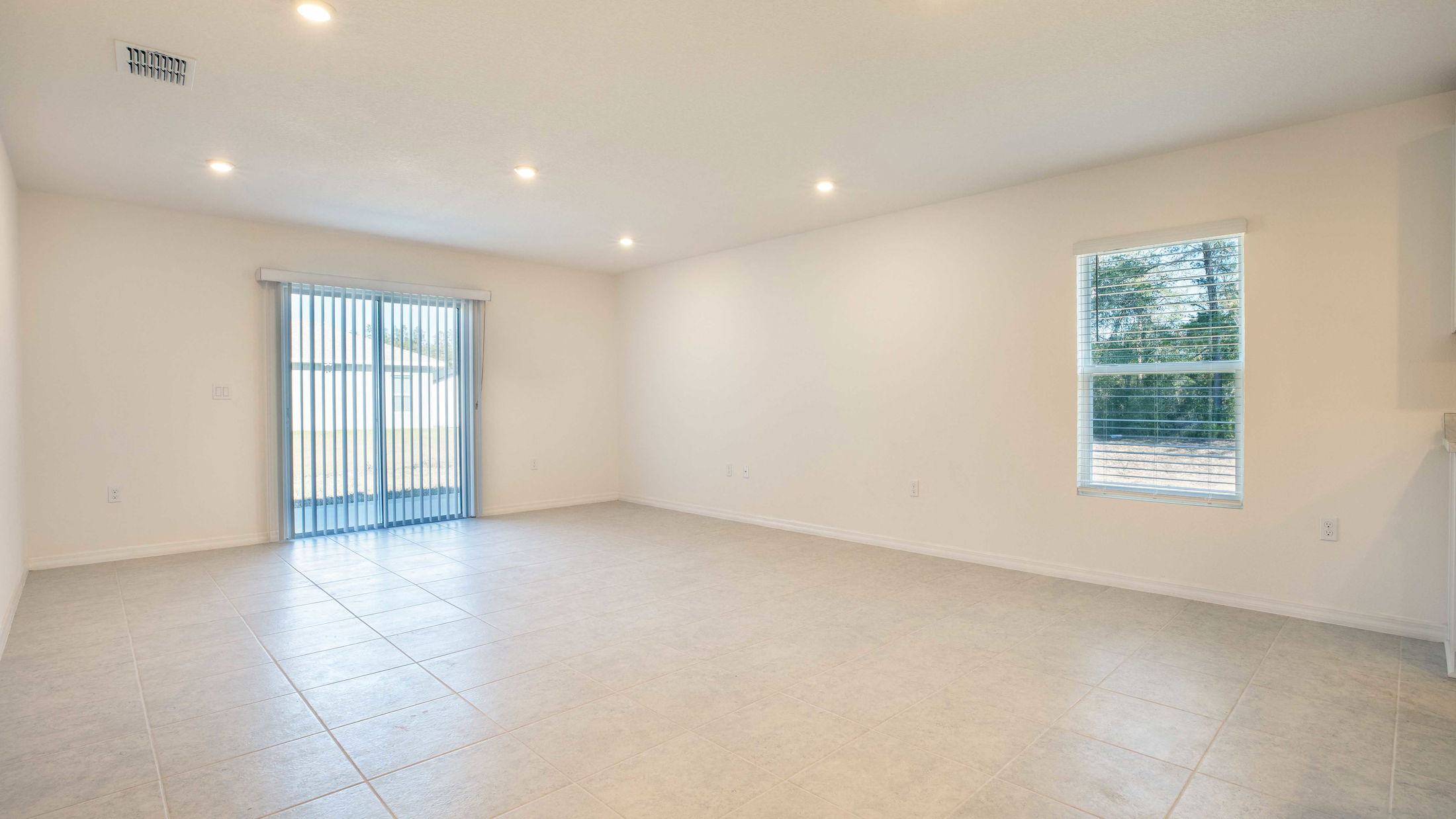 Wide-angle view of an empty, well-lit living room with large tile flooring, a window, and sliding glass doors with vertical blinds.